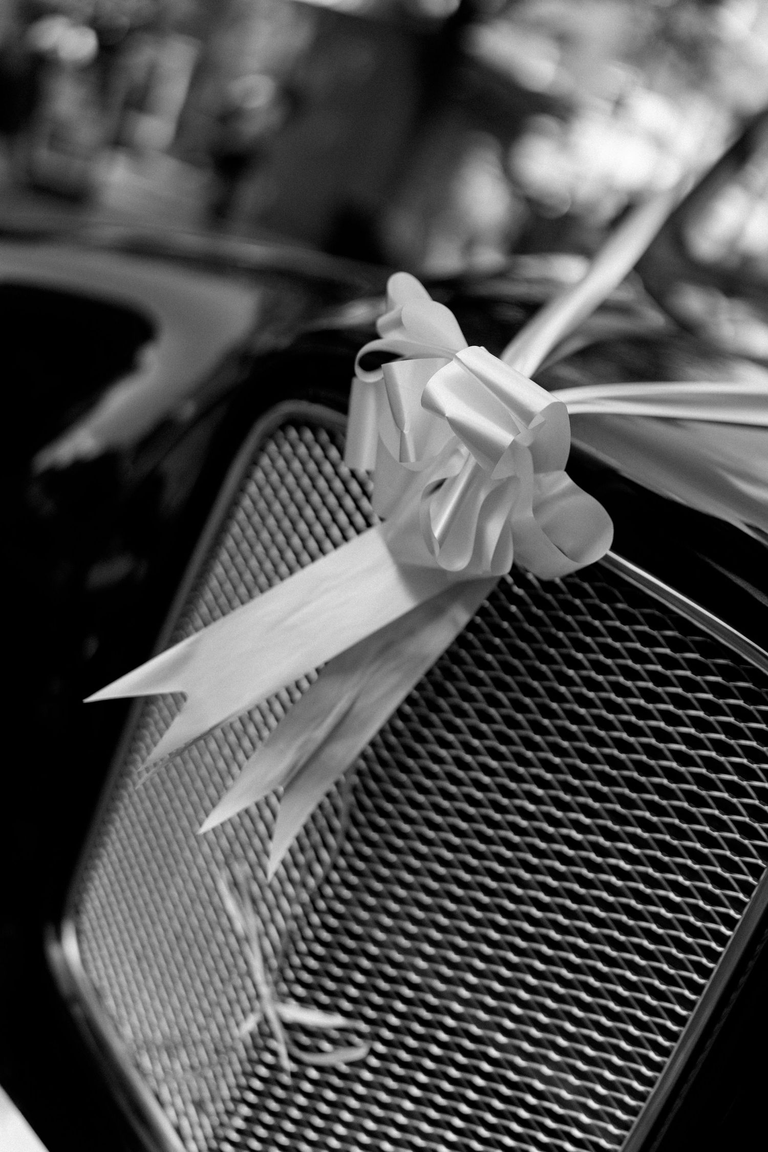 Black and white close-up of white satin ribbon bow on vintage wedding car grille.