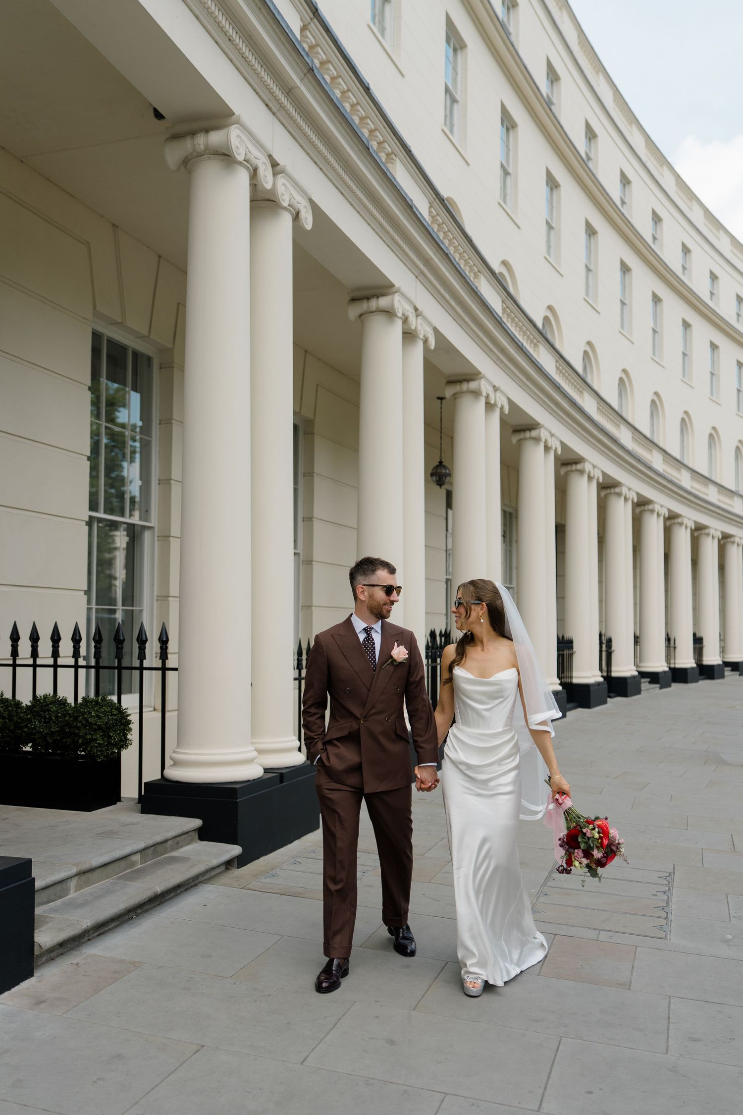 Elegant bride and groom walking hand in hand past curved white colonnade building on their wedding day.