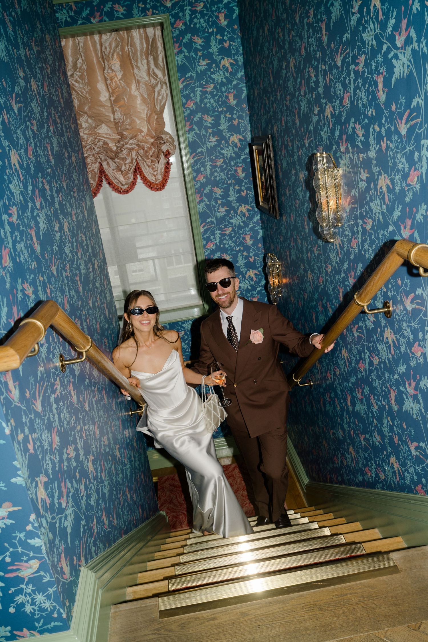 Bride and groom in sunglasses descend ornate staircase with blue floral wallpaper at a luxury wedding venue.