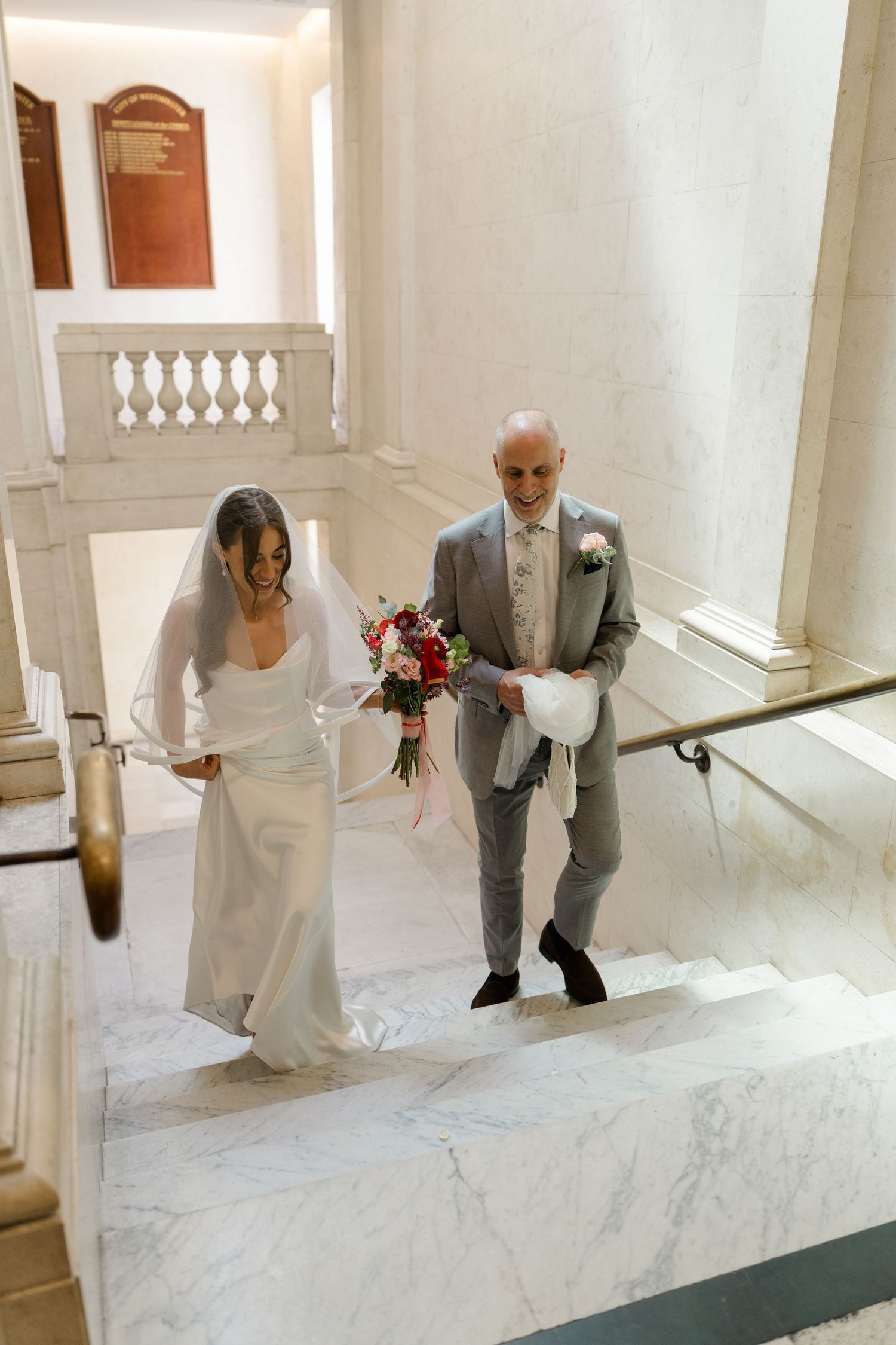 Bride in white gown and veil walks up marble stairs with older man in gray suit holding bouquet.