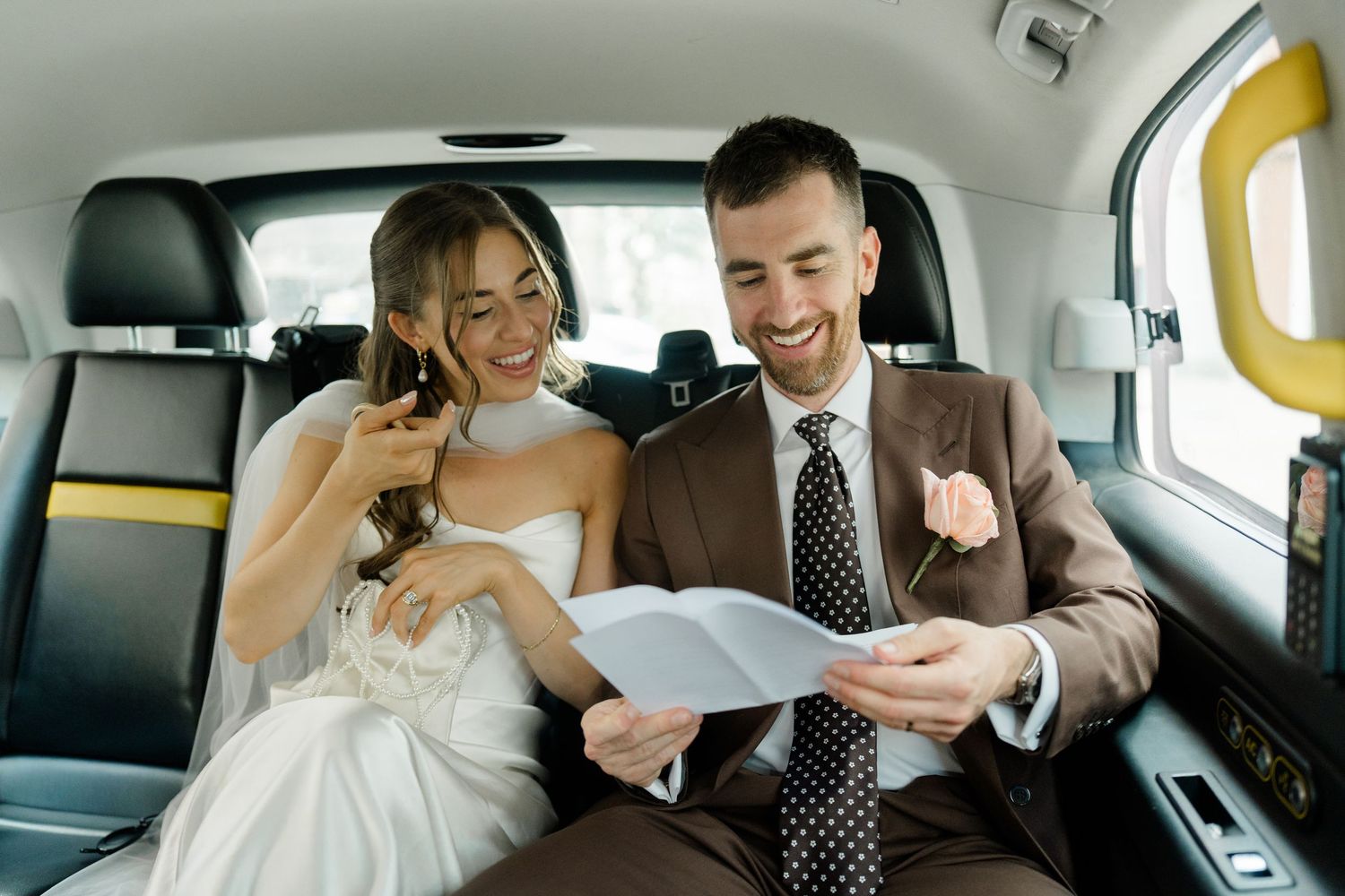 Happy bride and groom smile while reading a card together in the backseat of a black London taxi cab.
