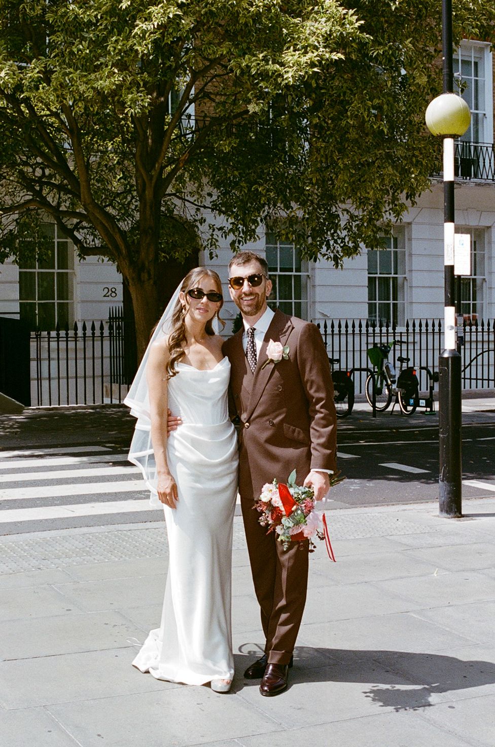 Bride and groom in sunglasses pose on sunny street corner after wedding ceremony.