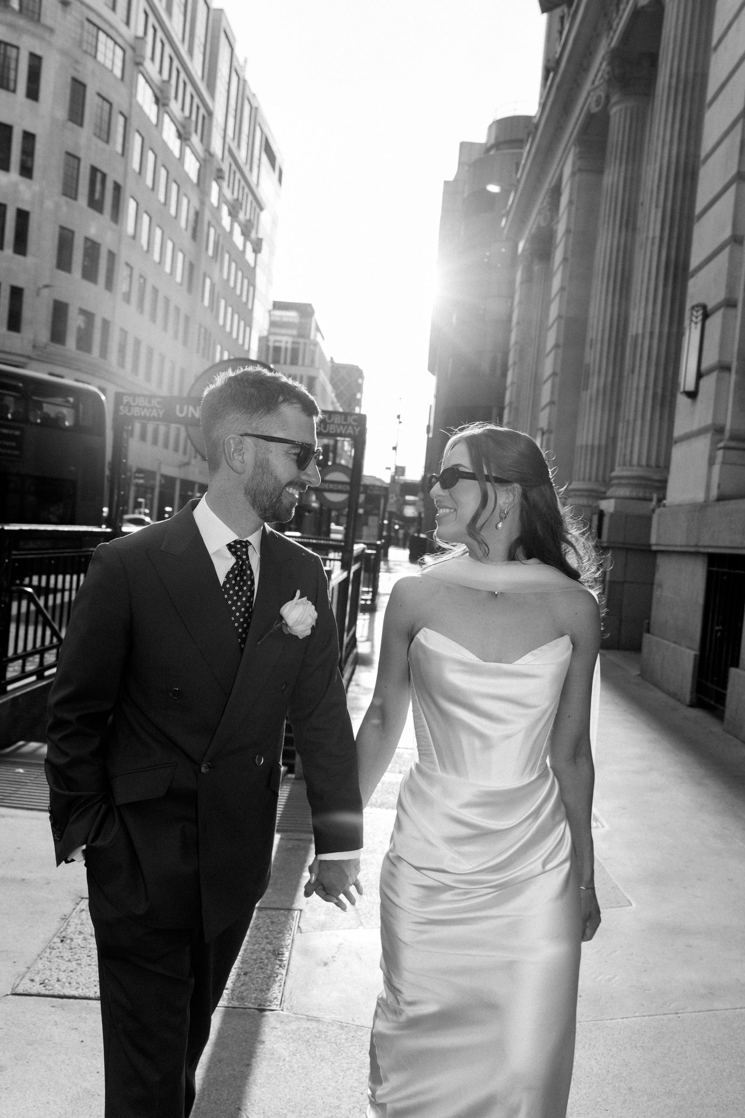 Black and white photo of bride and groom holding hands on a sunlit city street on their wedding day.