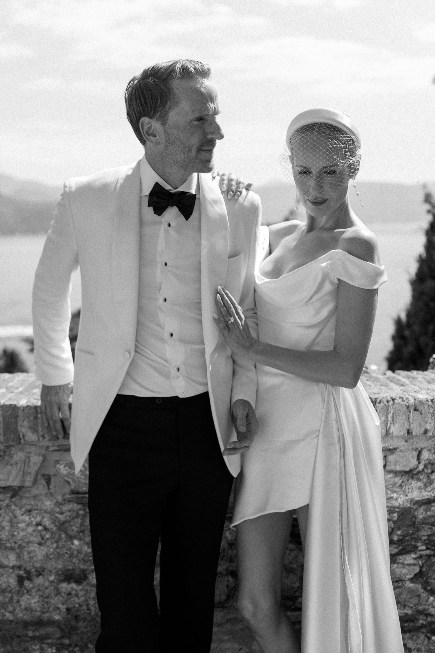 Elegant couple pose together, bride in off-shoulder mini dress with birdcage veil, groom in white tuxedo jacket.