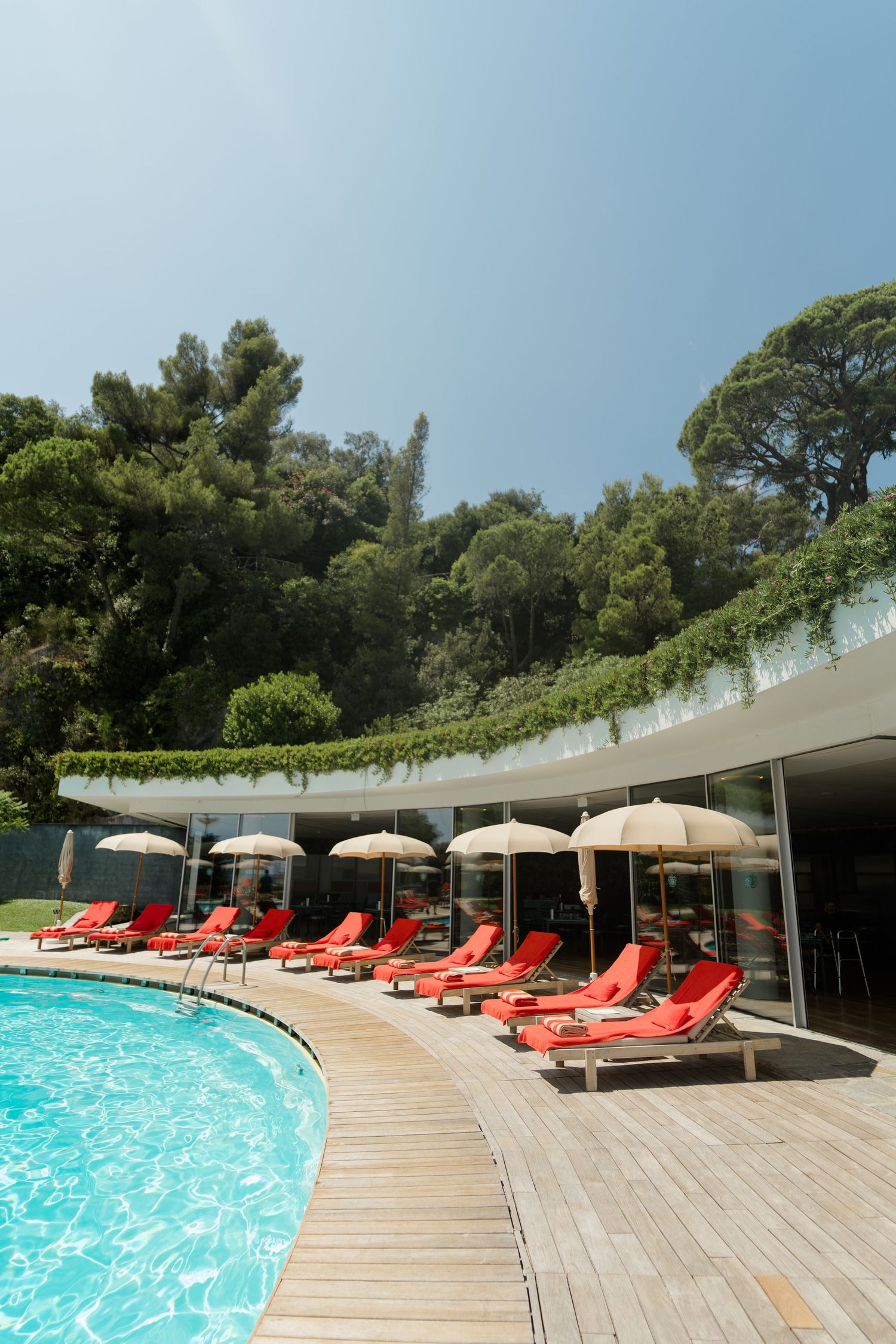 Luxury pool area with red lounge chairs, umbrellas, and lush green trees under a clear blue sky.