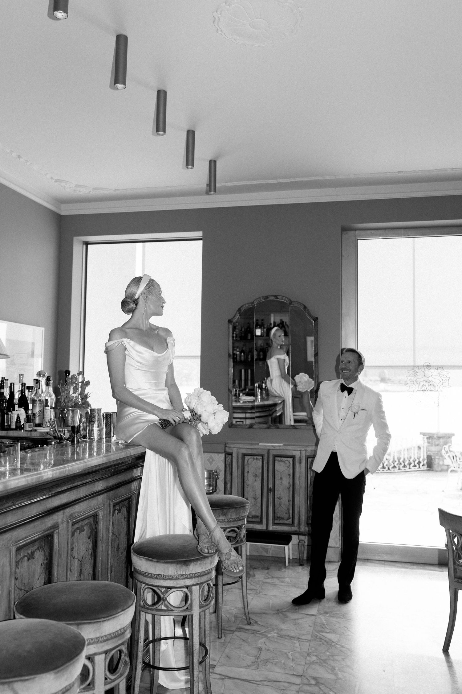 Bride sitting on bar counter holding bouquet while groom in white tuxedo stands nearby in black and white photo.