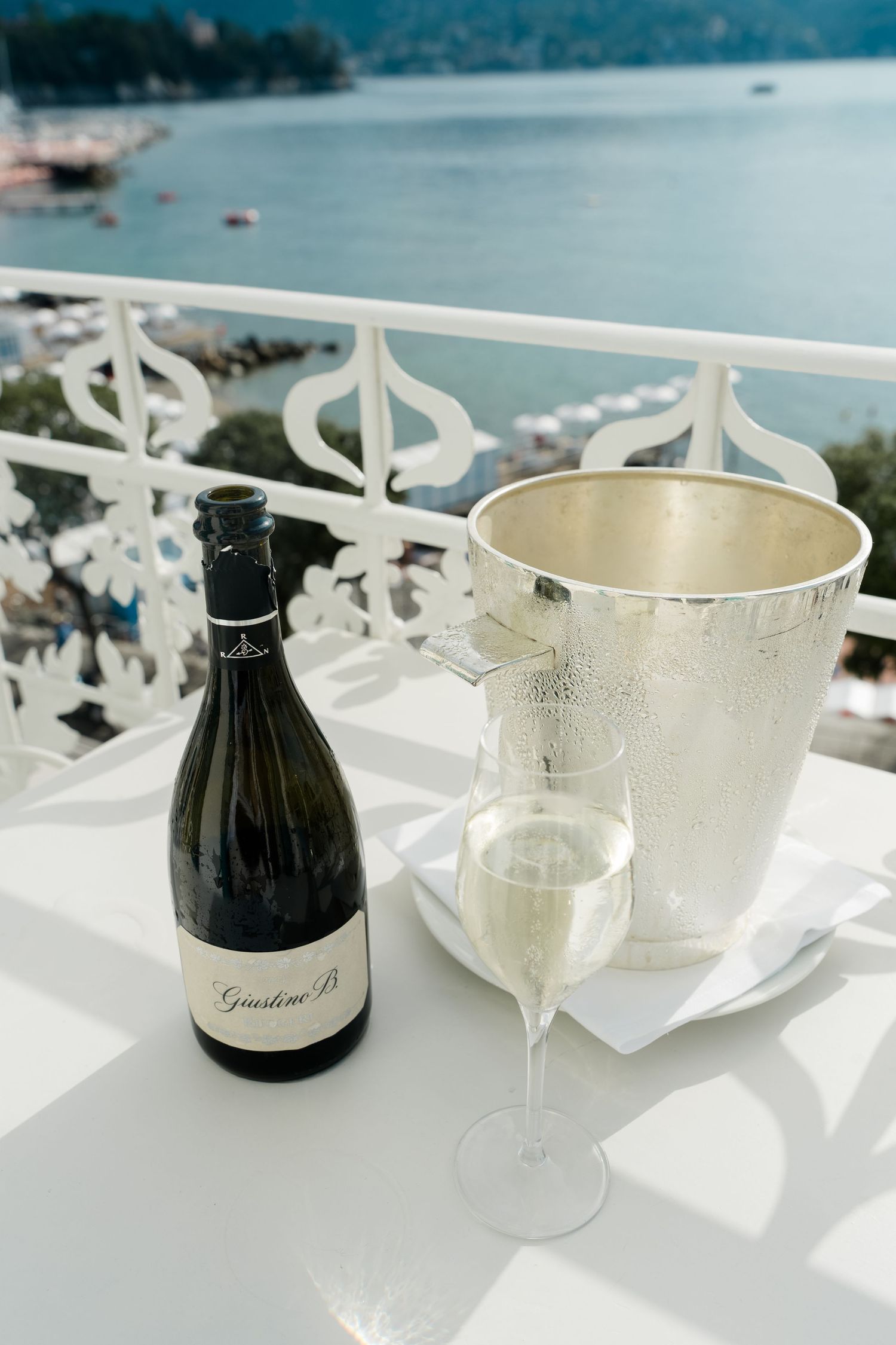 Bottle of Giustino B. Prosecco with a champagne flute and silver ice bucket on a white balcony table.