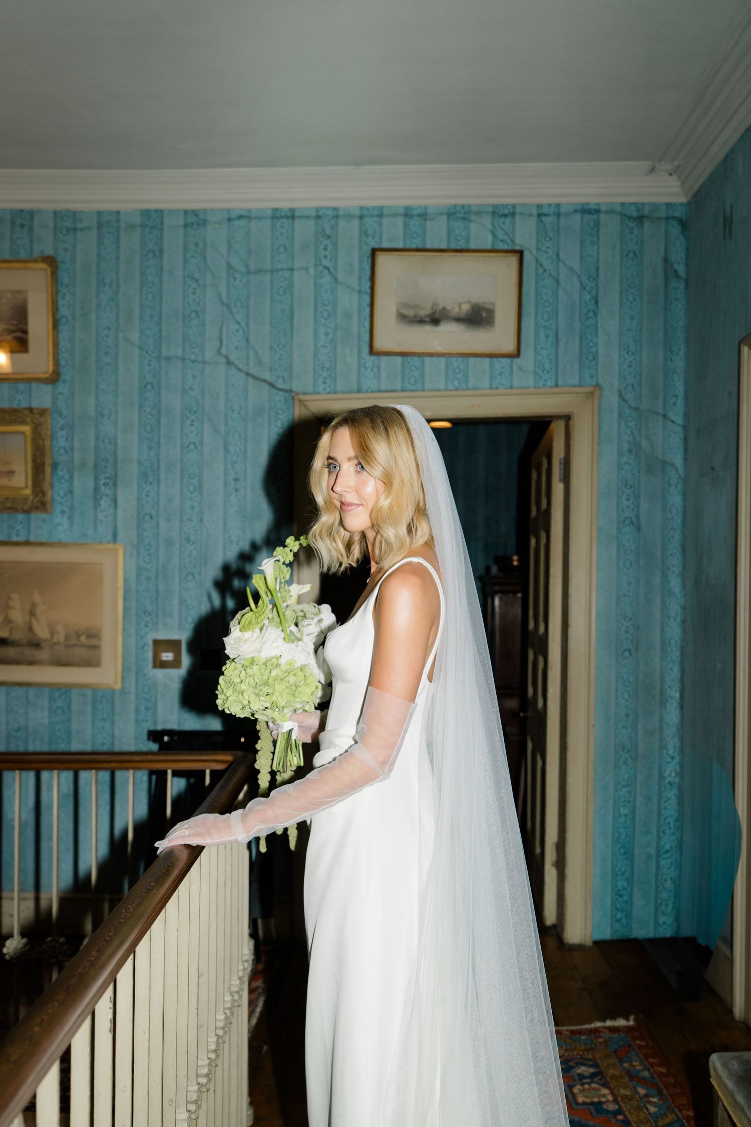 Bride in white gown and veil holds green bouquet on staircase with blue wallpaper.