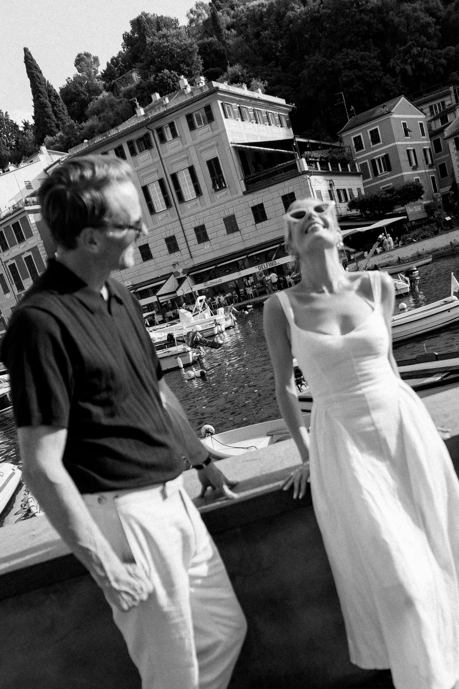Black and white photo of a couple near a Mediterranean harbor, woman in white dress, man in dark shirt, boats and buildings behind.