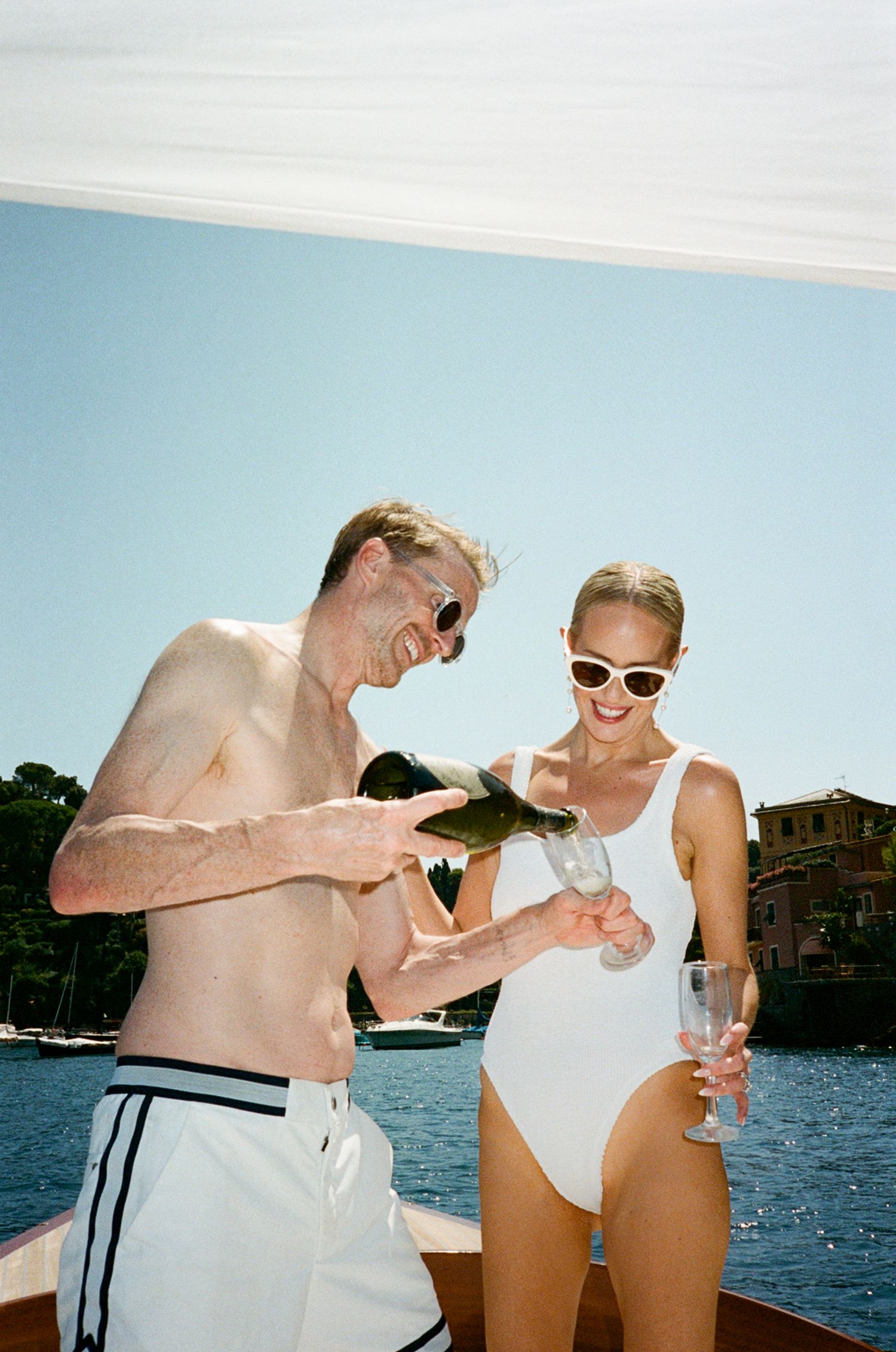Couple on a boat in swimwear, pouring champagne and laughing, with blue water and coastline in the background.