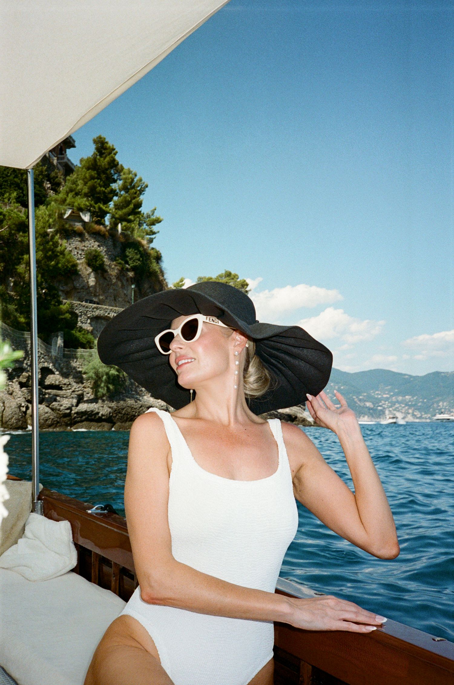 Elegant blonde woman in white swimsuit and large black sun hat on a boat in Italy.