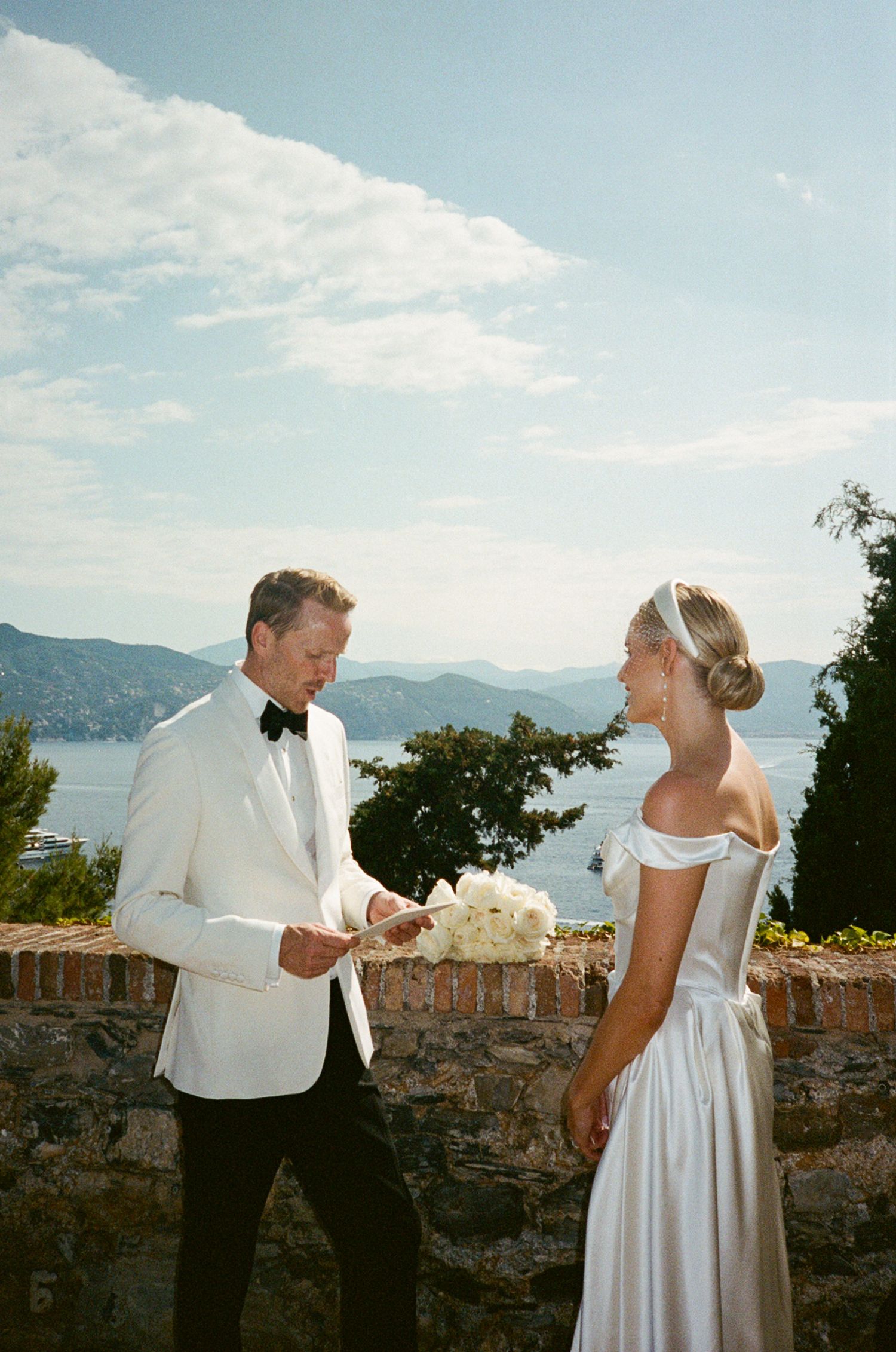 Elegant groom in white tuxedo presenting bouquet to bride in off-shoulder gown with scenic lake backdrop.