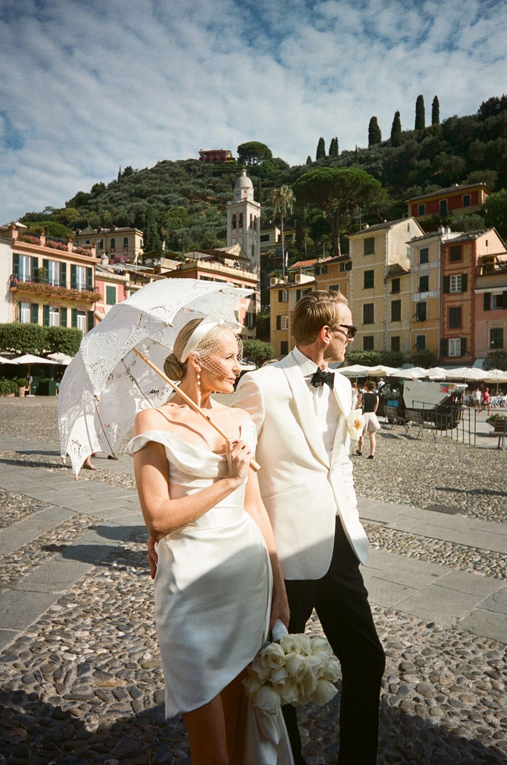 Elegant bride in short white dress with parasol and groom in white tuxedo stroll through Italian piazza.