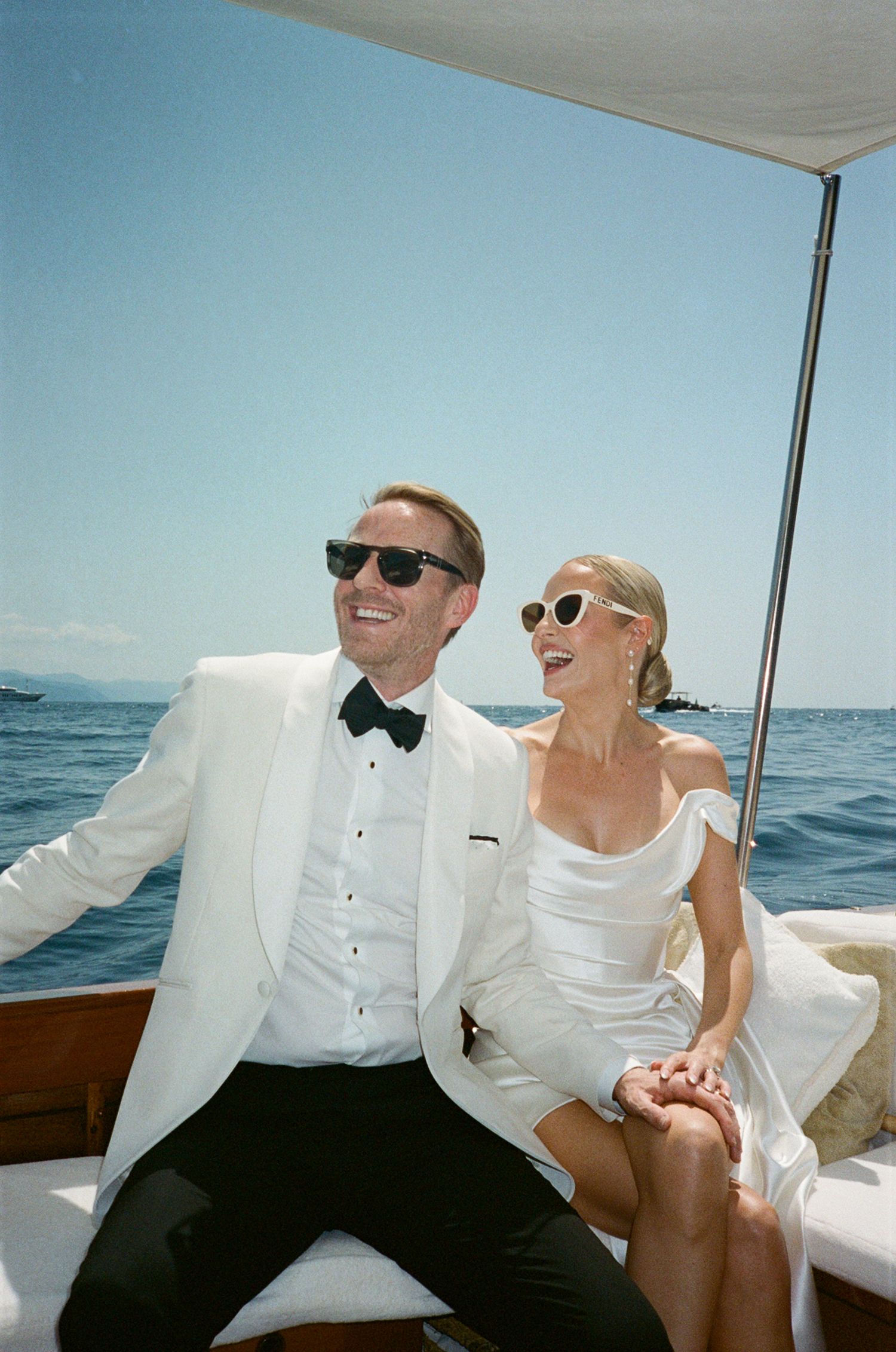 Newlyweds in white wedding attire laugh together on luxury boat with ocean backdrop.