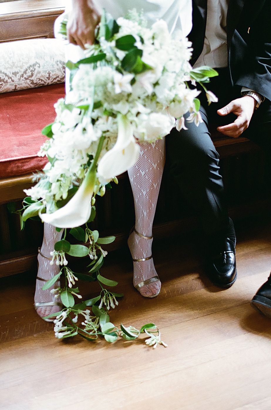 Close-up view of formal black shoes and white dress hem with cascading floral bouquet on hardwood floor.