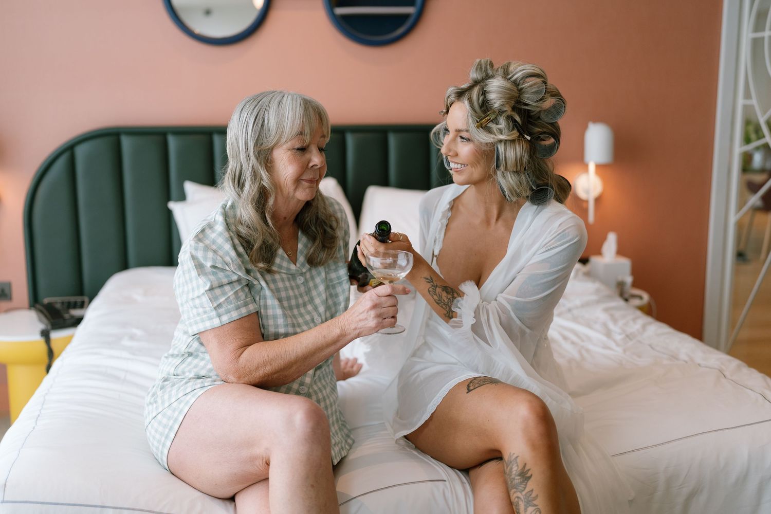 Two people sitting on a bed sharing drinks during wedding preparations.