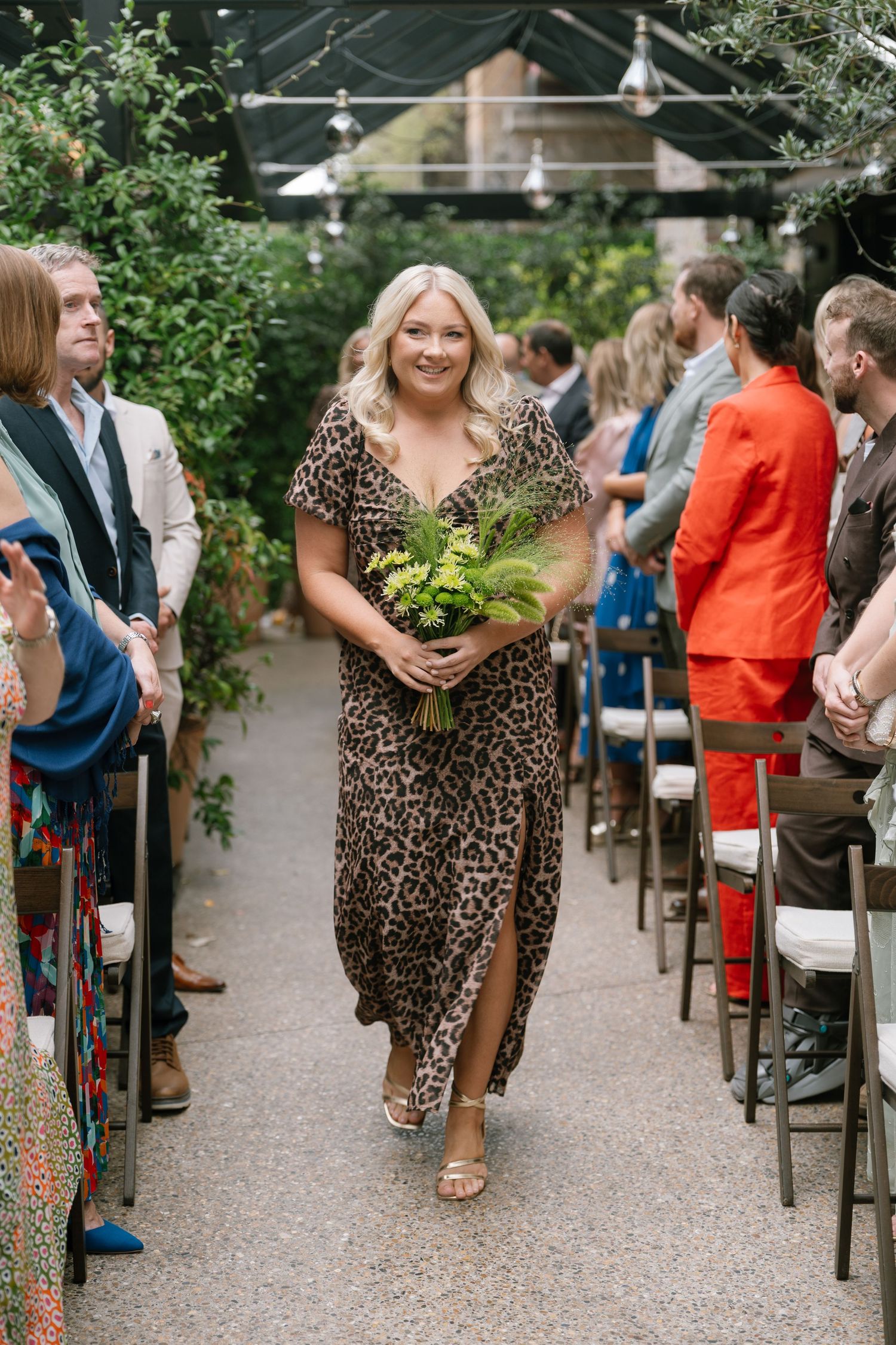 Wedding guest in leopard print dress walks down aisle holding greenery at outdoor ceremony.