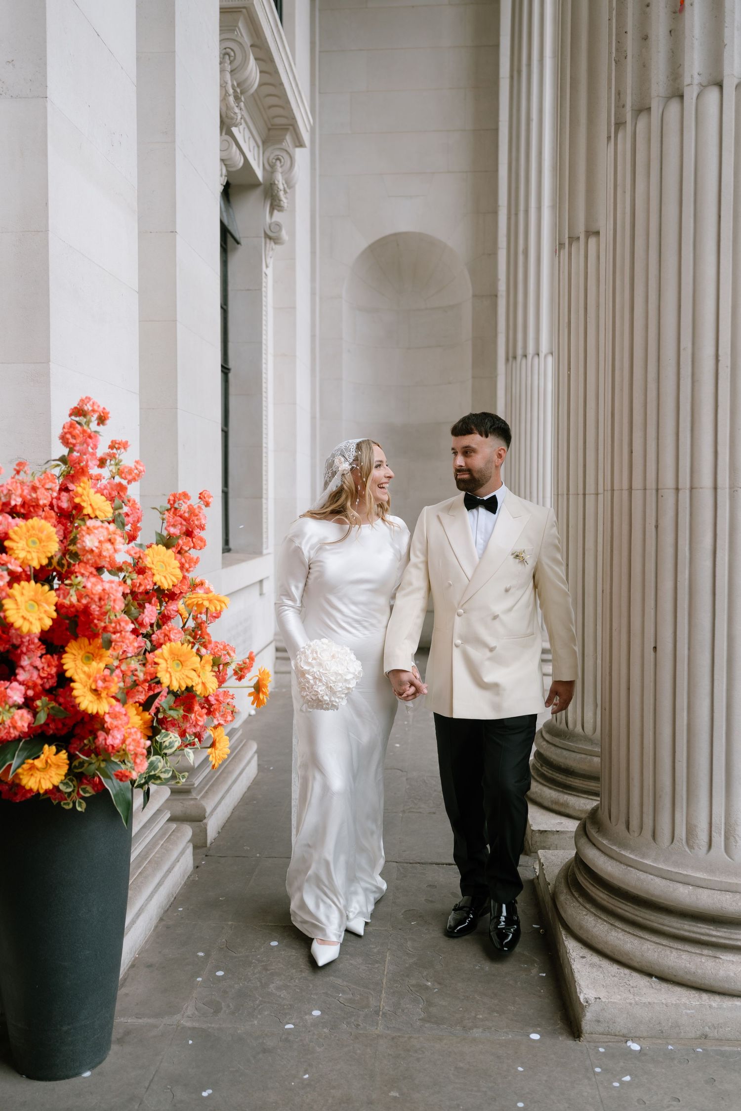 Wedding couple walking among classical white columns with orange floral arrangements.