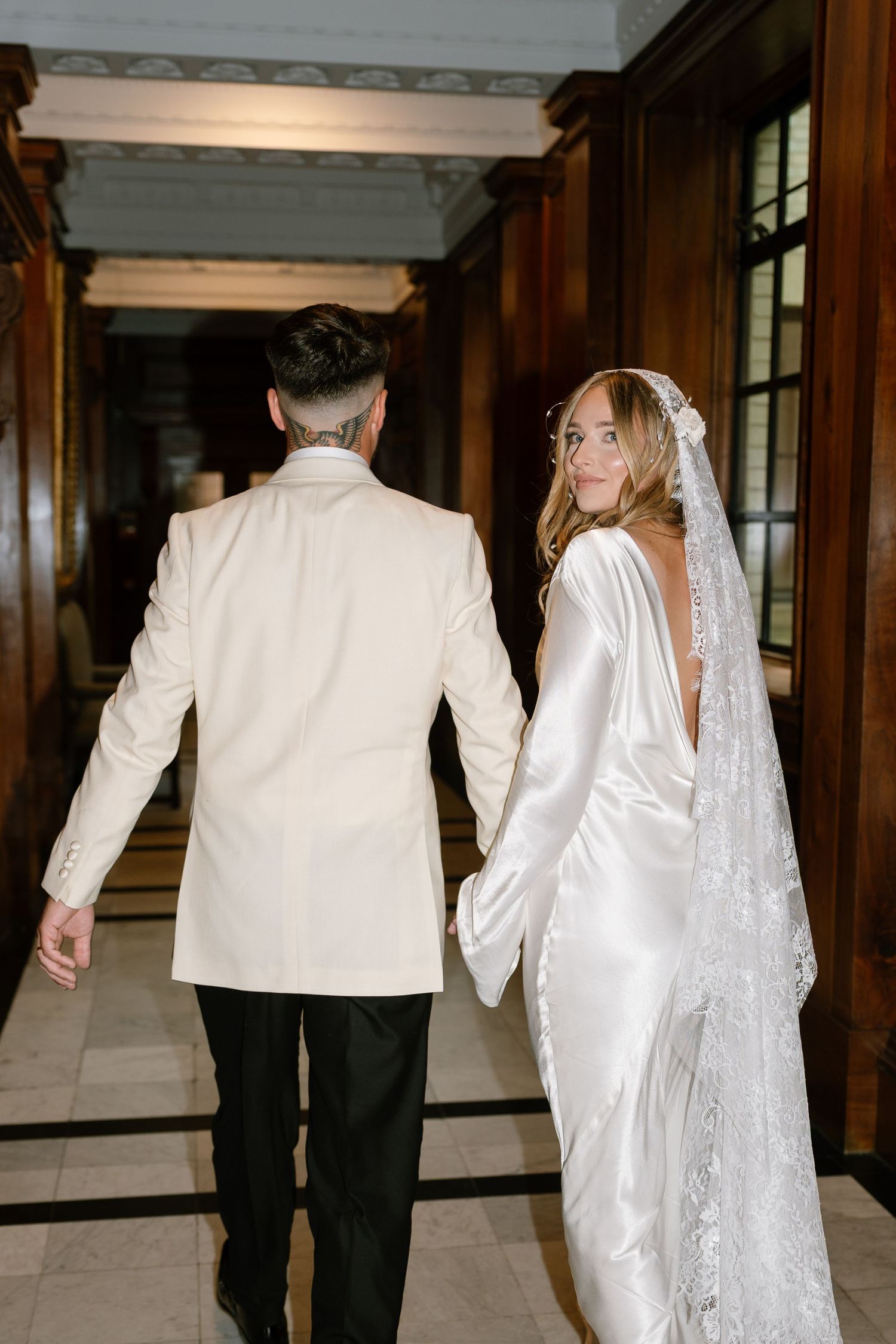 Wedding couple walking down an elegant wood-paneled hallway in formal attire.