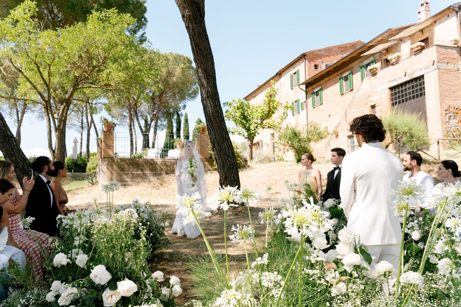 Outdoor wedding ceremony setup with white floral arrangements and tall trees.