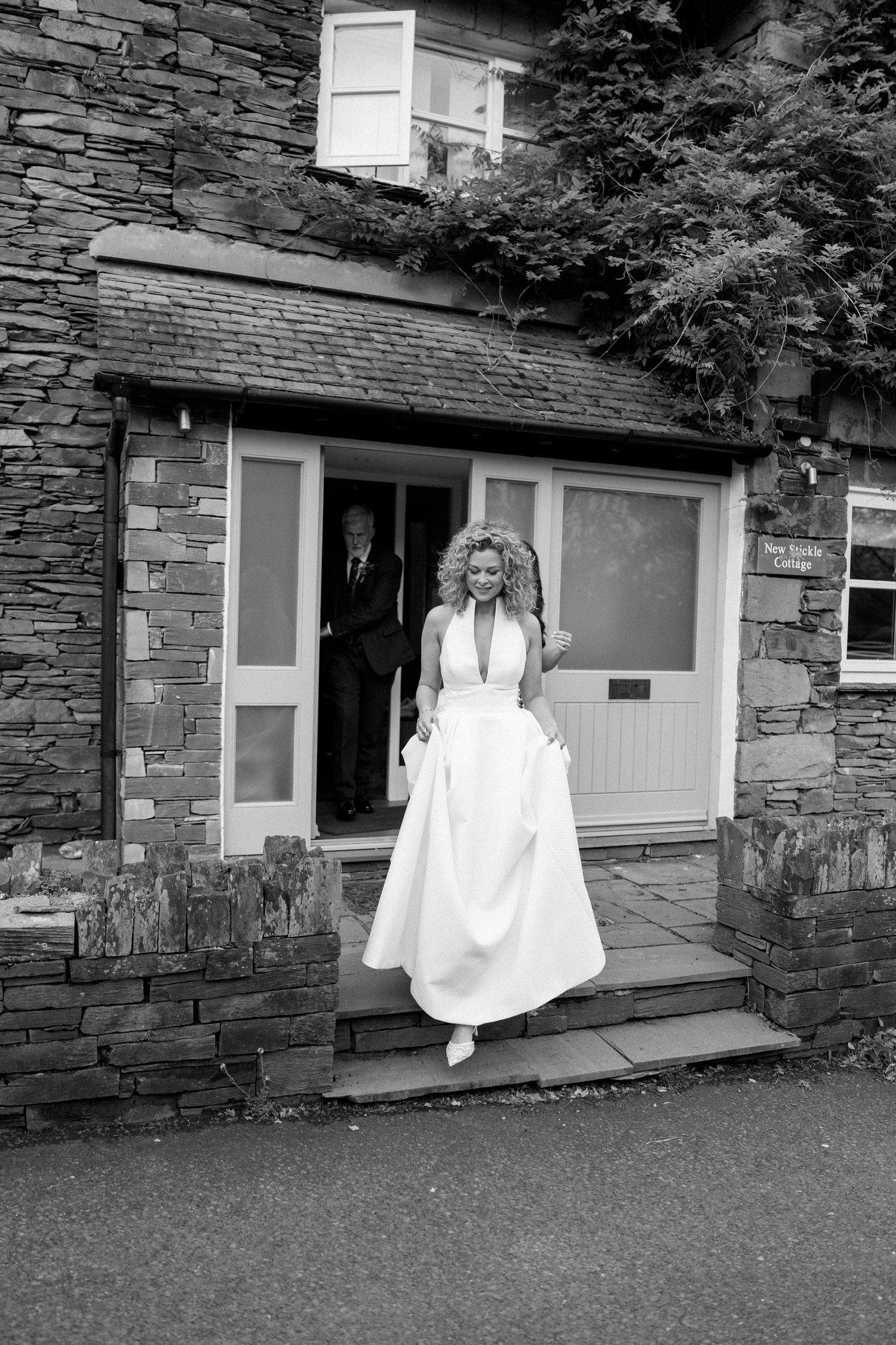 A wedding scene outside a rustic stone cottage with a white-dressed figure standing on the steps.