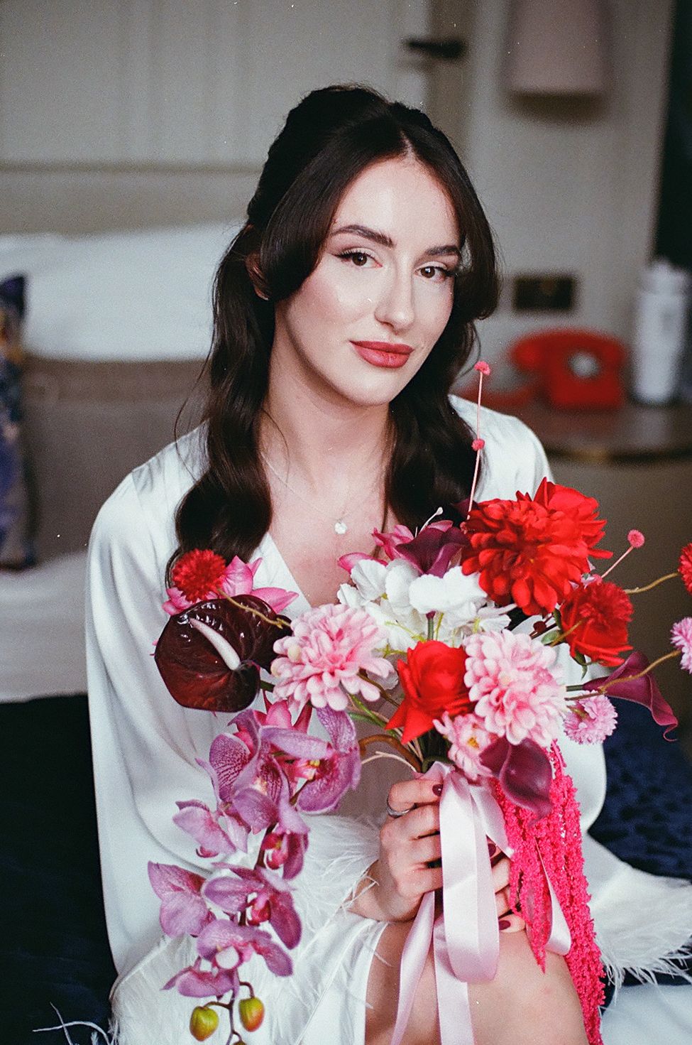 Woman in white holding vibrant pink and red flowers against a blurred interior background.