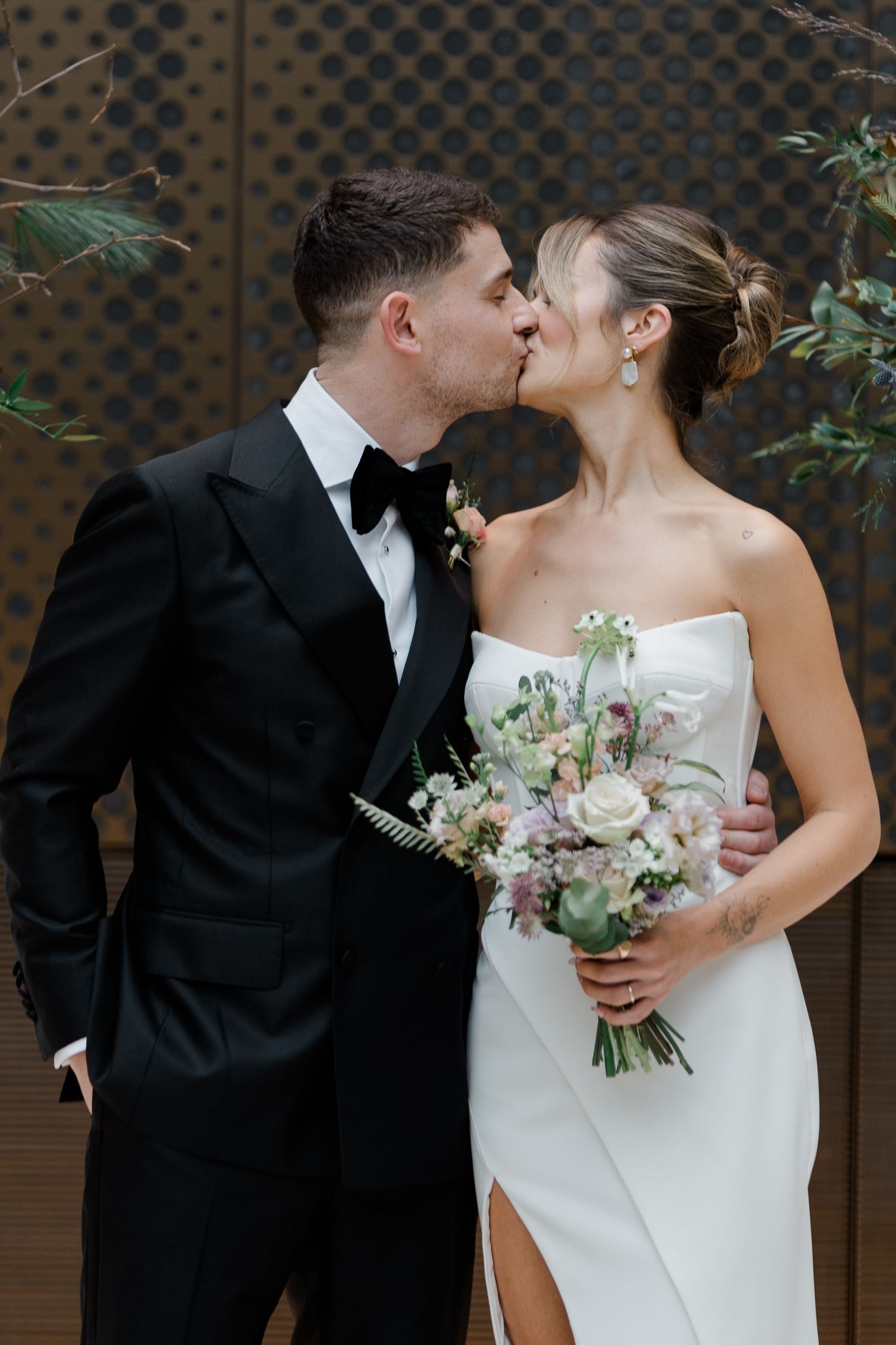 A couple shares romantic kisses in their wedding attire against a dark textured wall while holding a white floral bouquet.