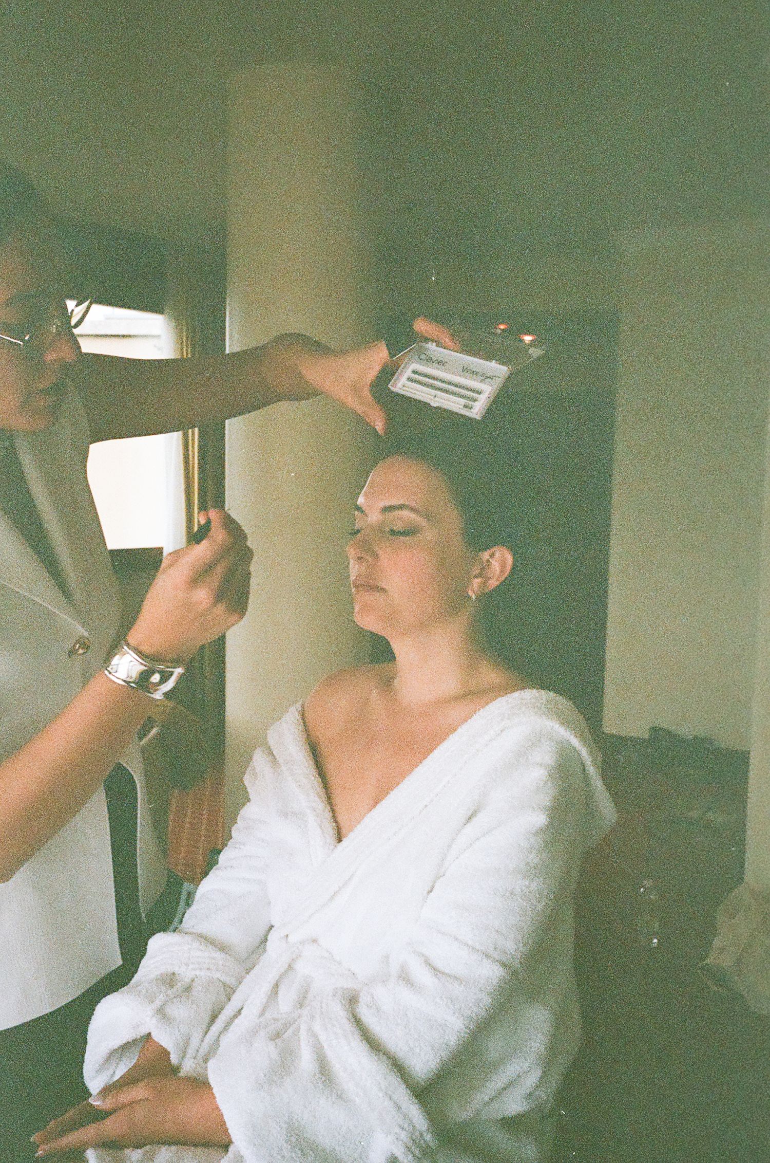Getting ready scene with bride in white robe having hair styled.