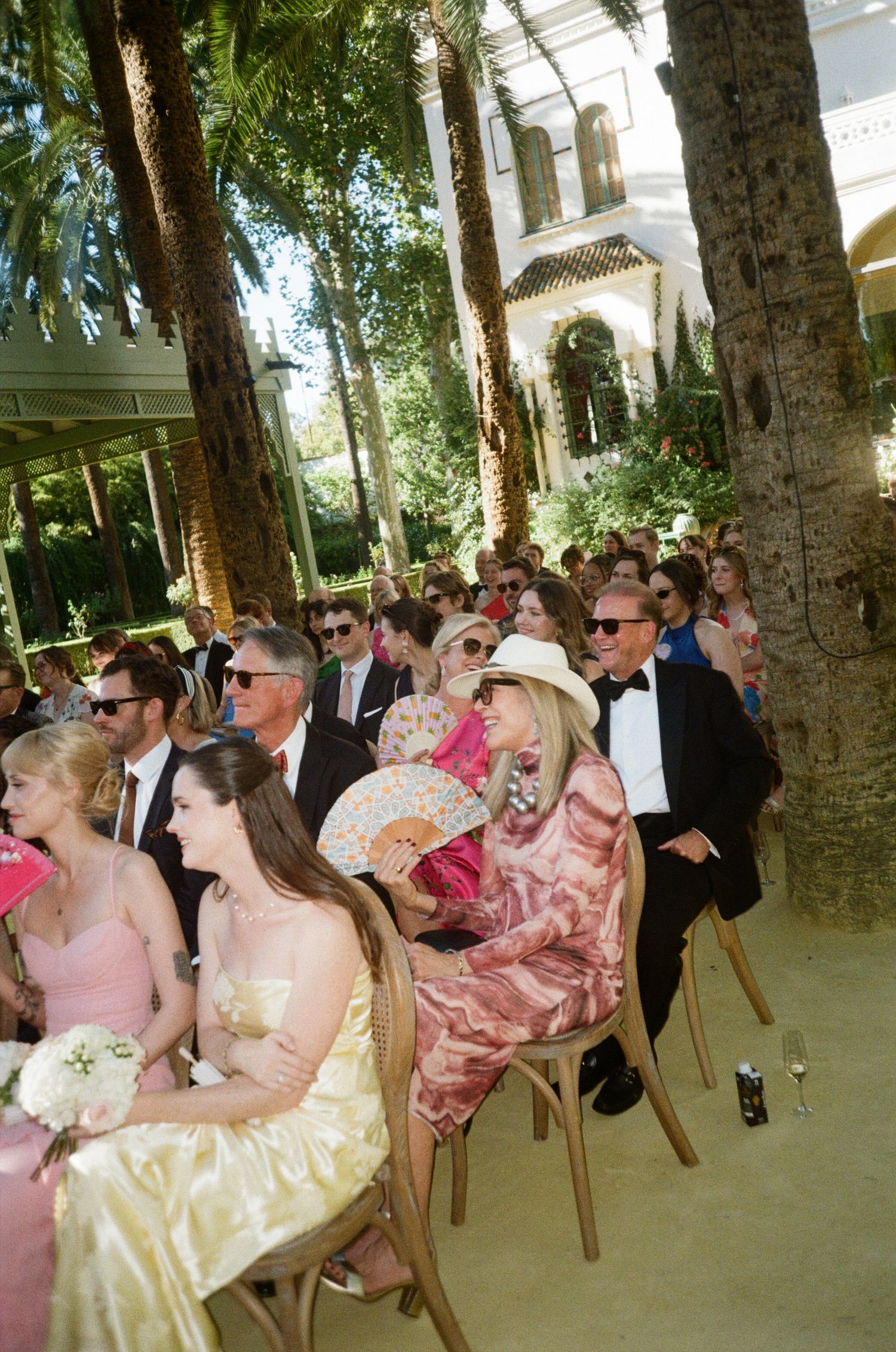 Wedding guests seated outdoors under palm trees at a formal garden ceremony.