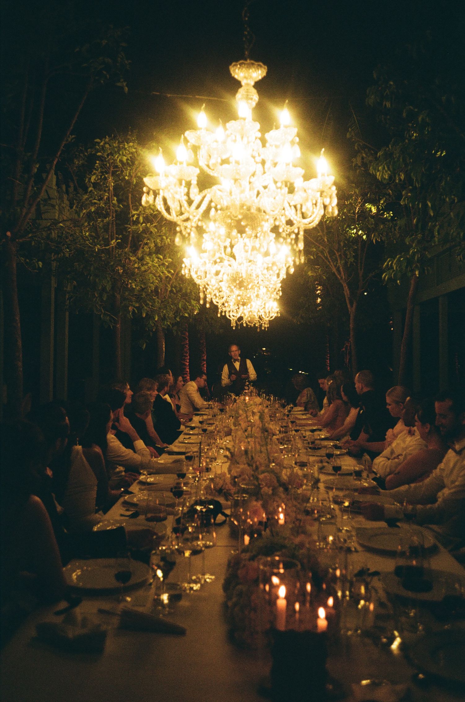 An intimate outdoor dinner setting illuminated by glowing crystal chandeliers with guests seated at a long table.
