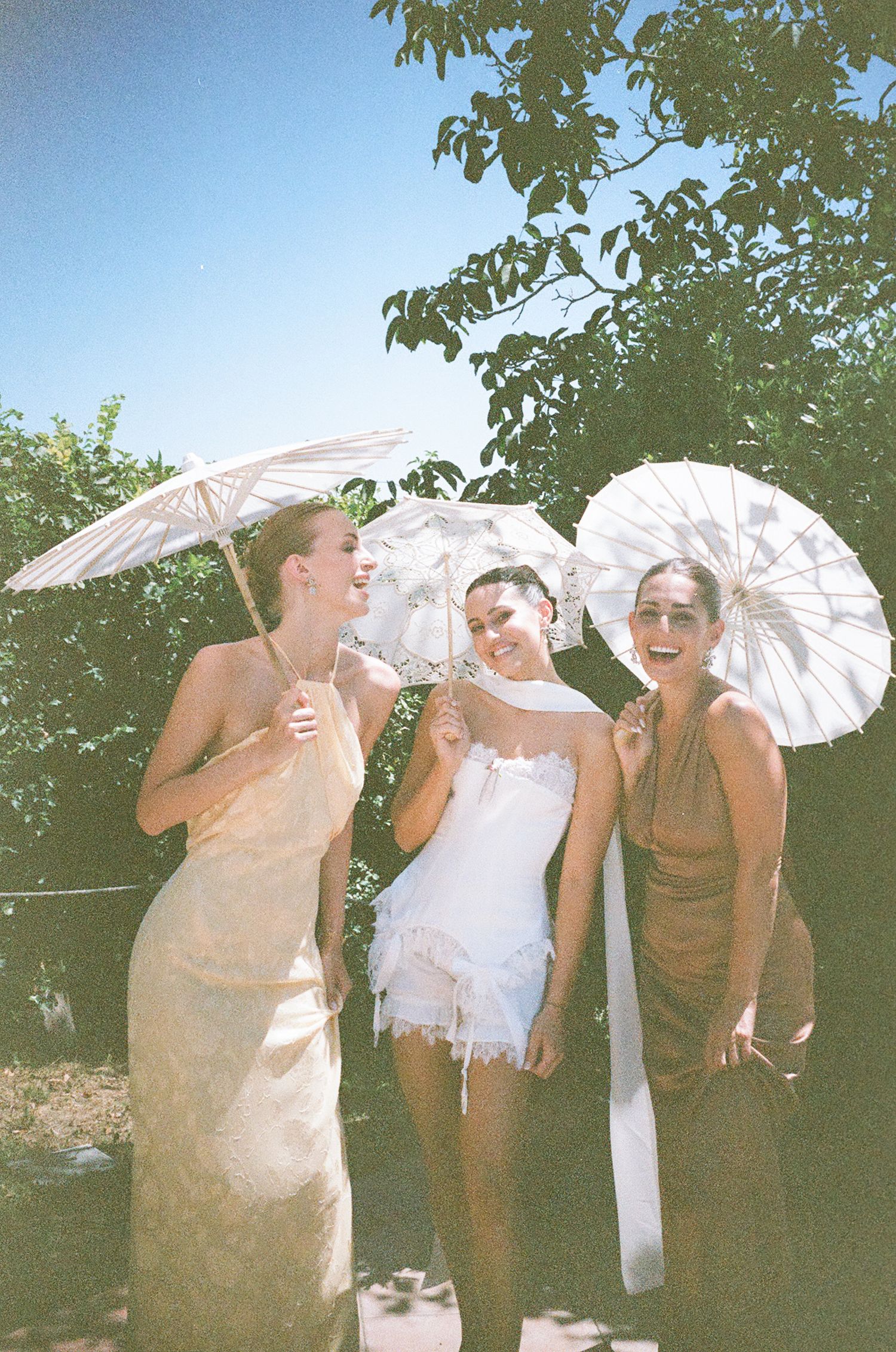 Three people pose playfully with white parasols in a sunny outdoor garden setting.