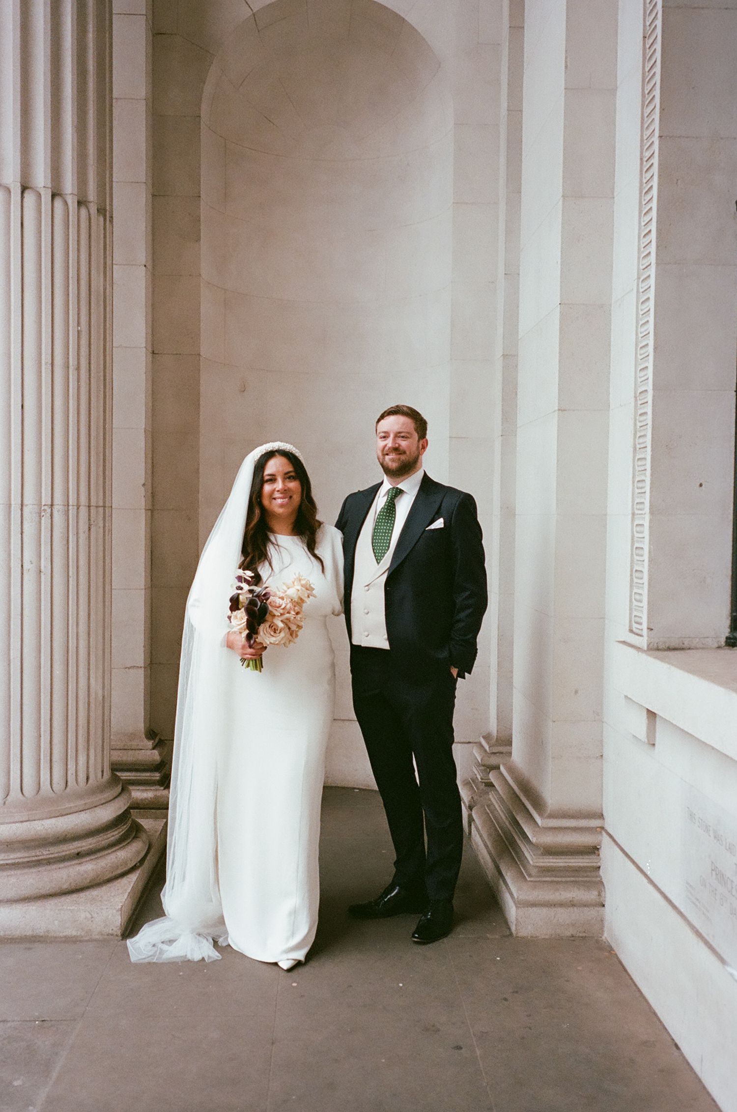 A bride and groom pose together against white columns in an elegant architectural setting for their wedding portraits.