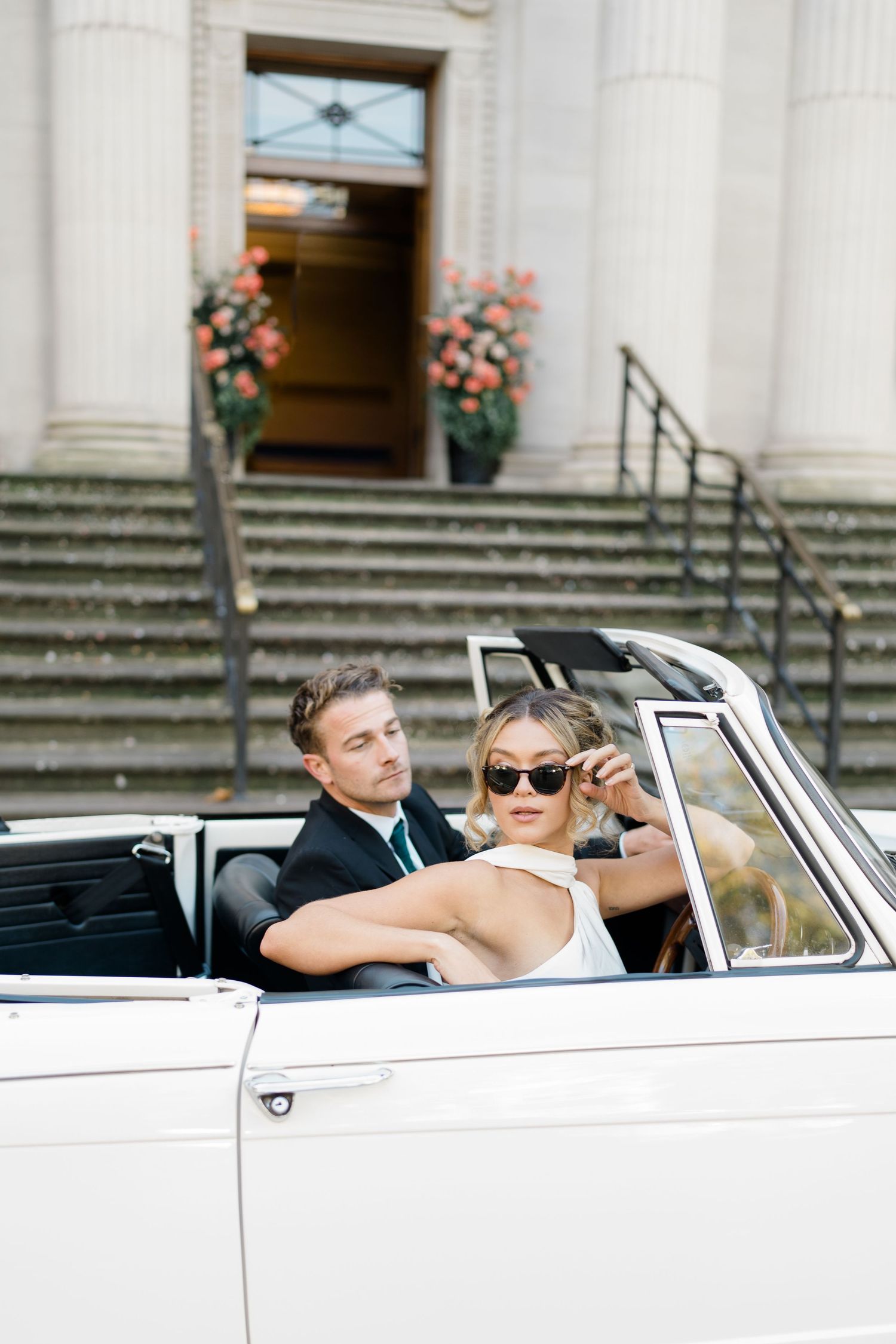 Couple poses playfully in a classic white convertible car in front of a grand building with stone steps.