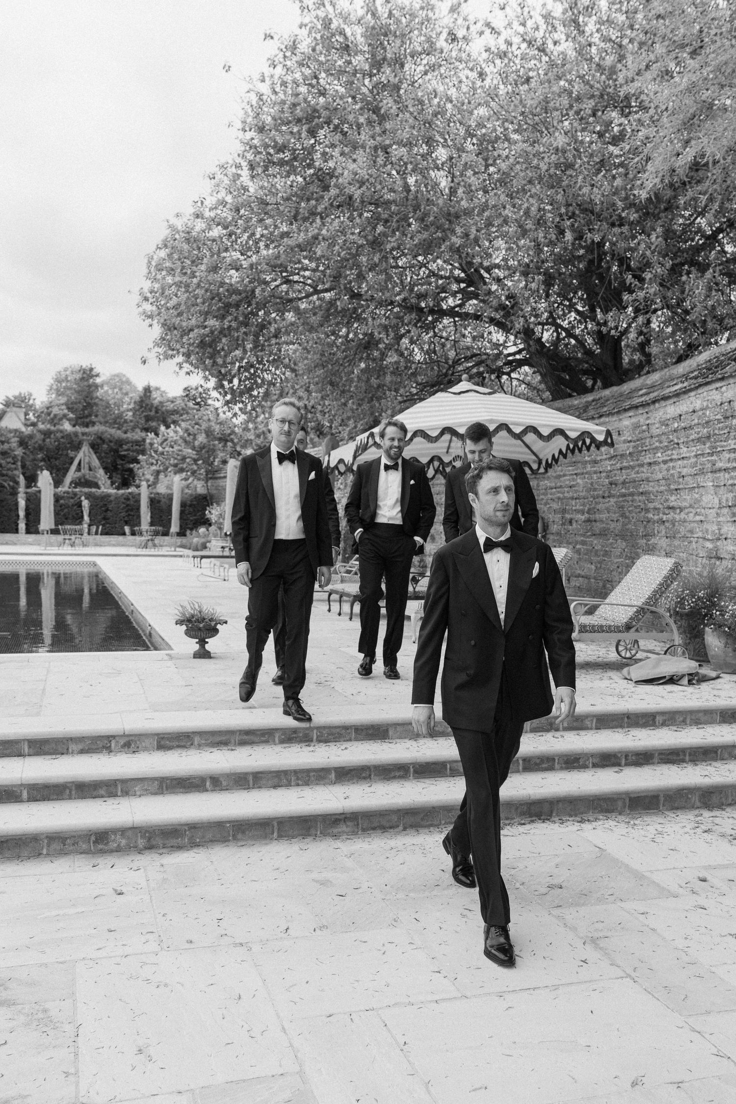 Groomsmen in black tuxedos walking by a pool at an elegant outdoor wedding venue.