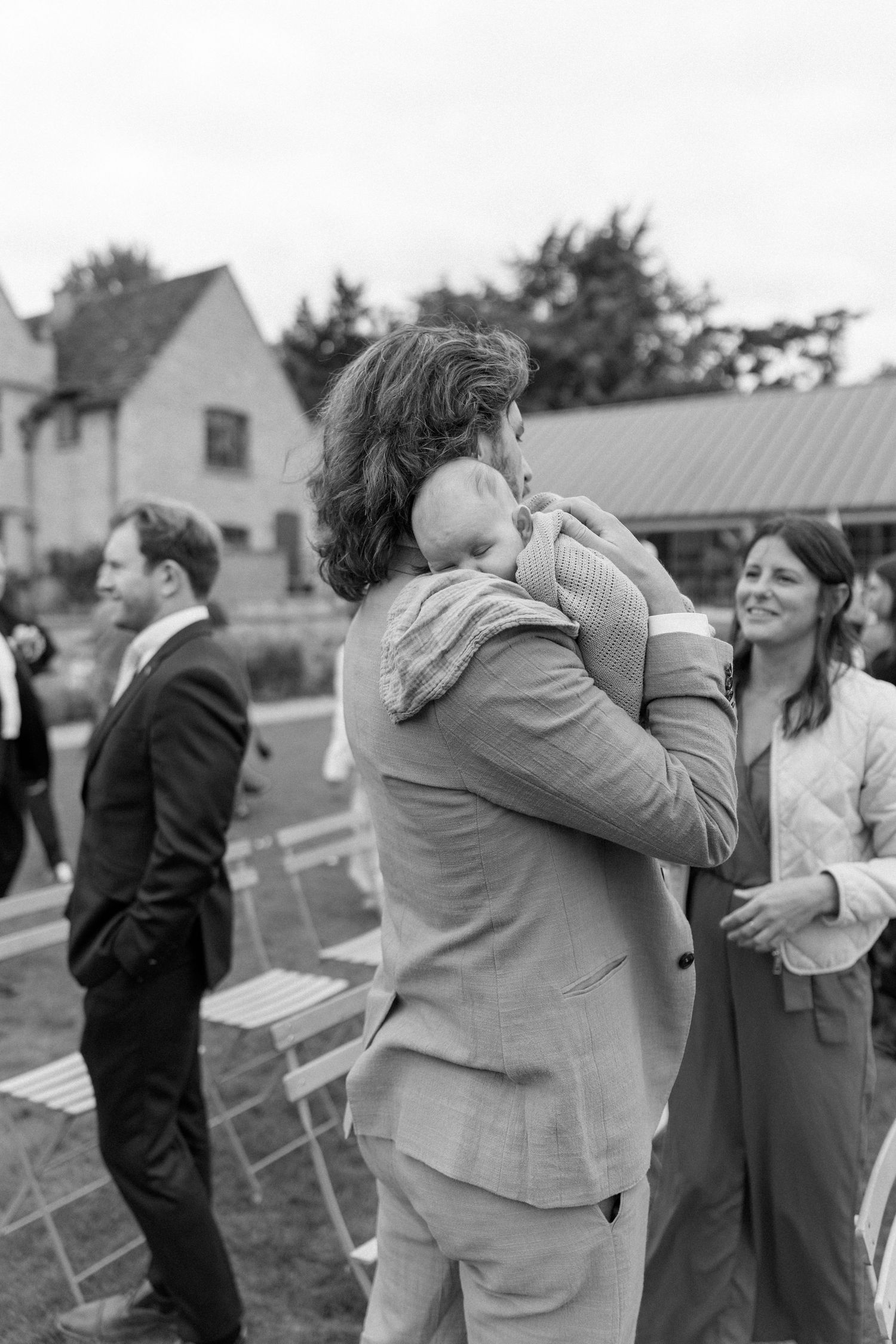 Wedding guest in grey suit holds sleeping baby during outdoor reception in black and white photo.
