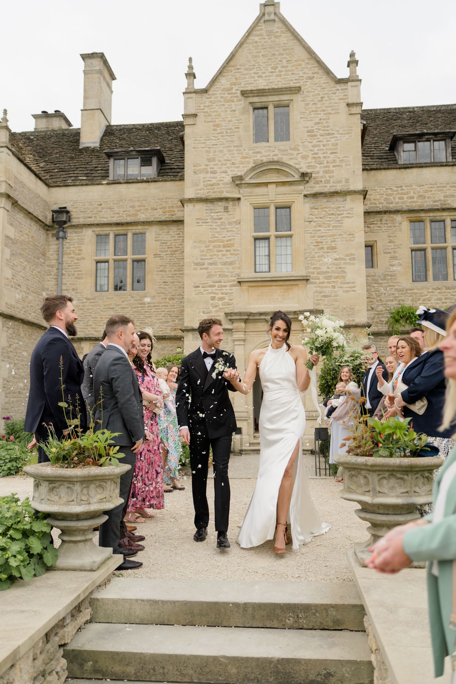 Bride and groom walk down steps as guests throw confetti outside a grand stone manor house wedding venue.