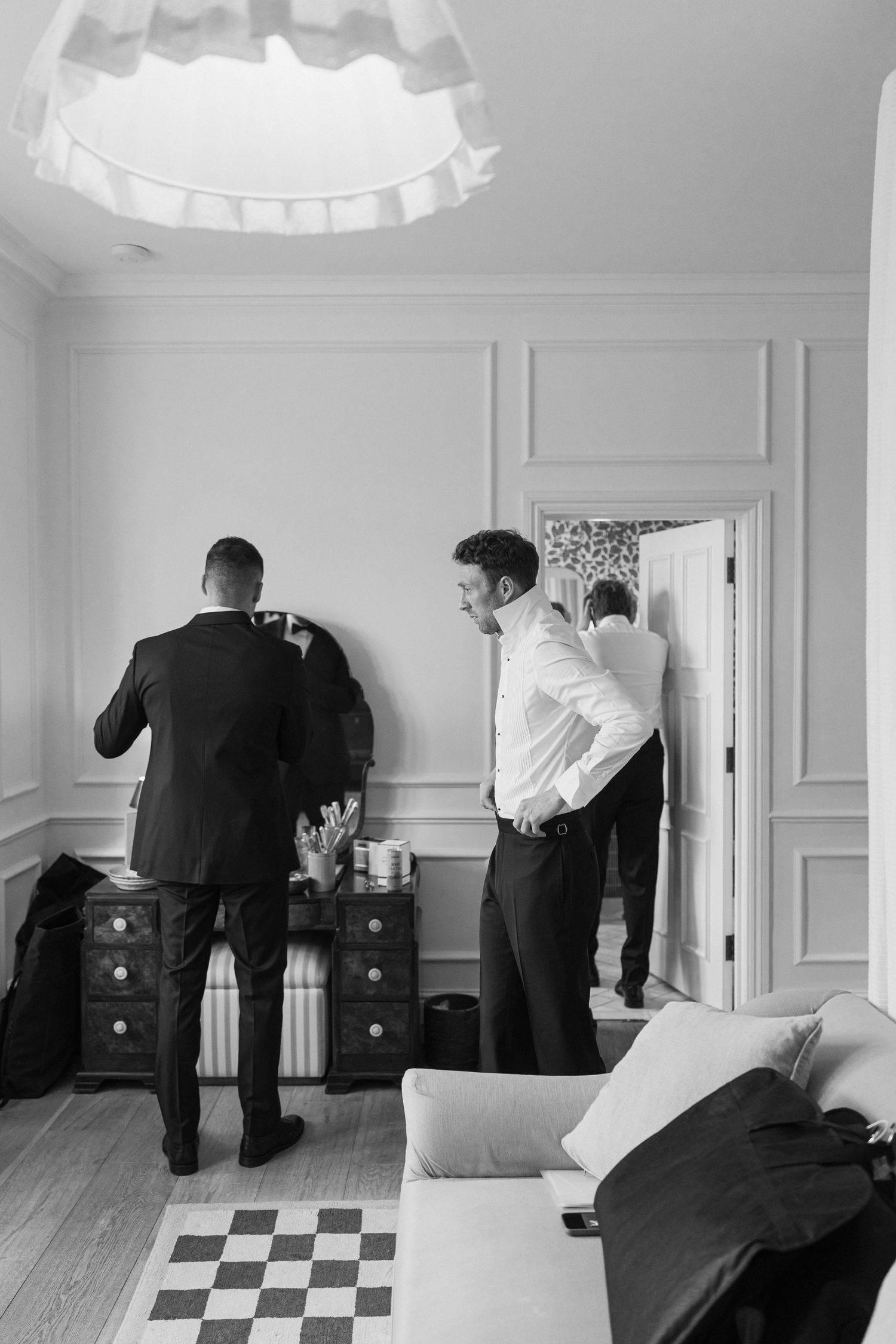 Two groomsmen getting dressed in formal black suits in a classic white-paneled hotel room.