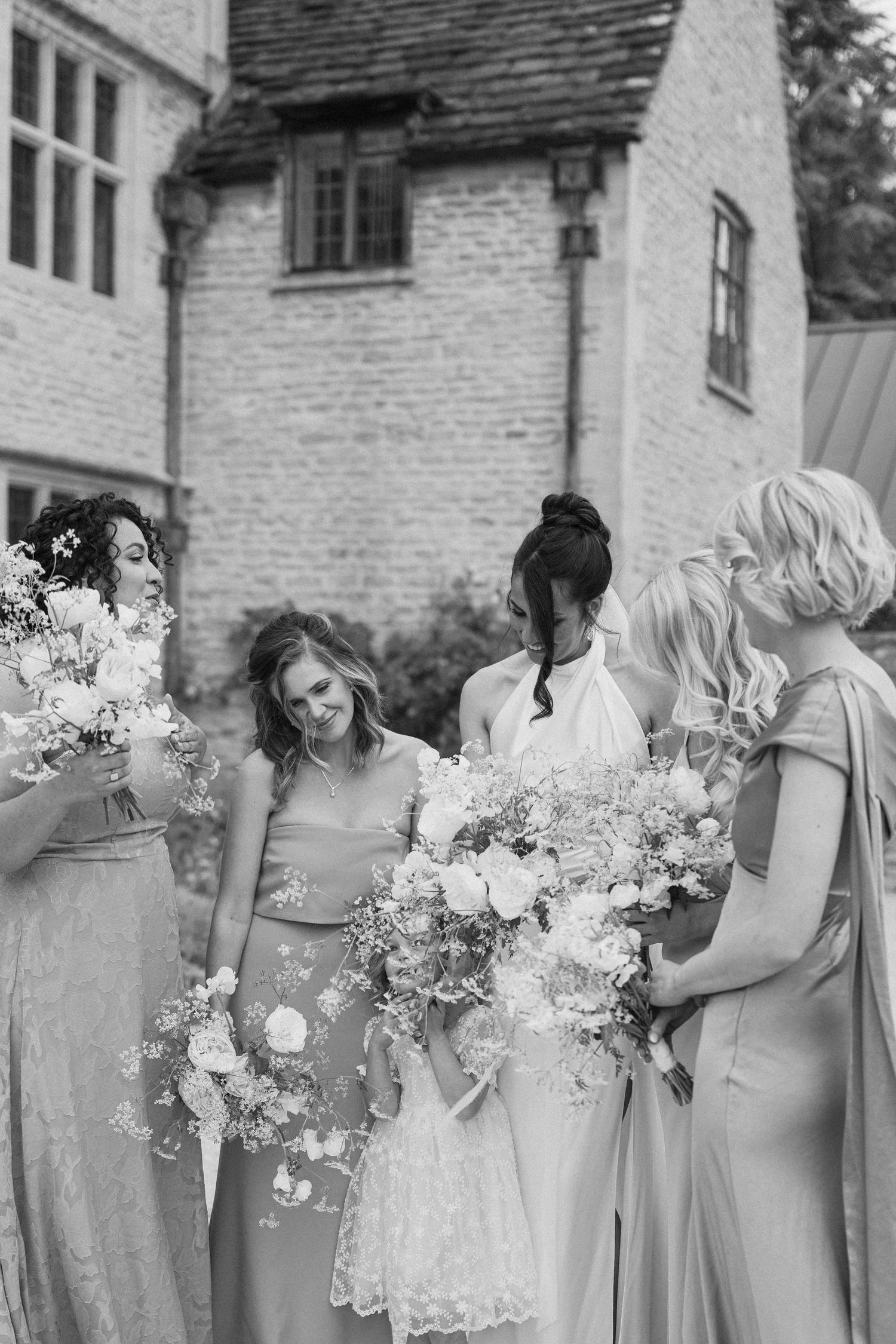 Black and white photo of bride and bridesmaids holding white floral bouquets outside stone venue.