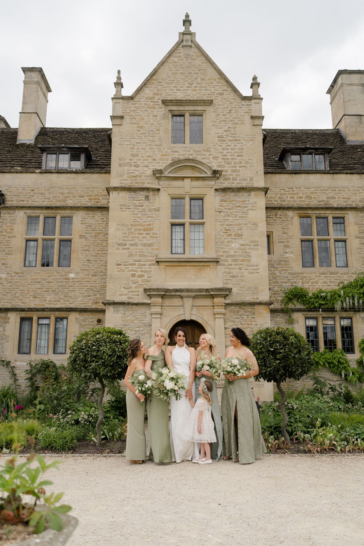 Bride and bridesmaids in sage green gowns pose outside a grand English stone manor house.
