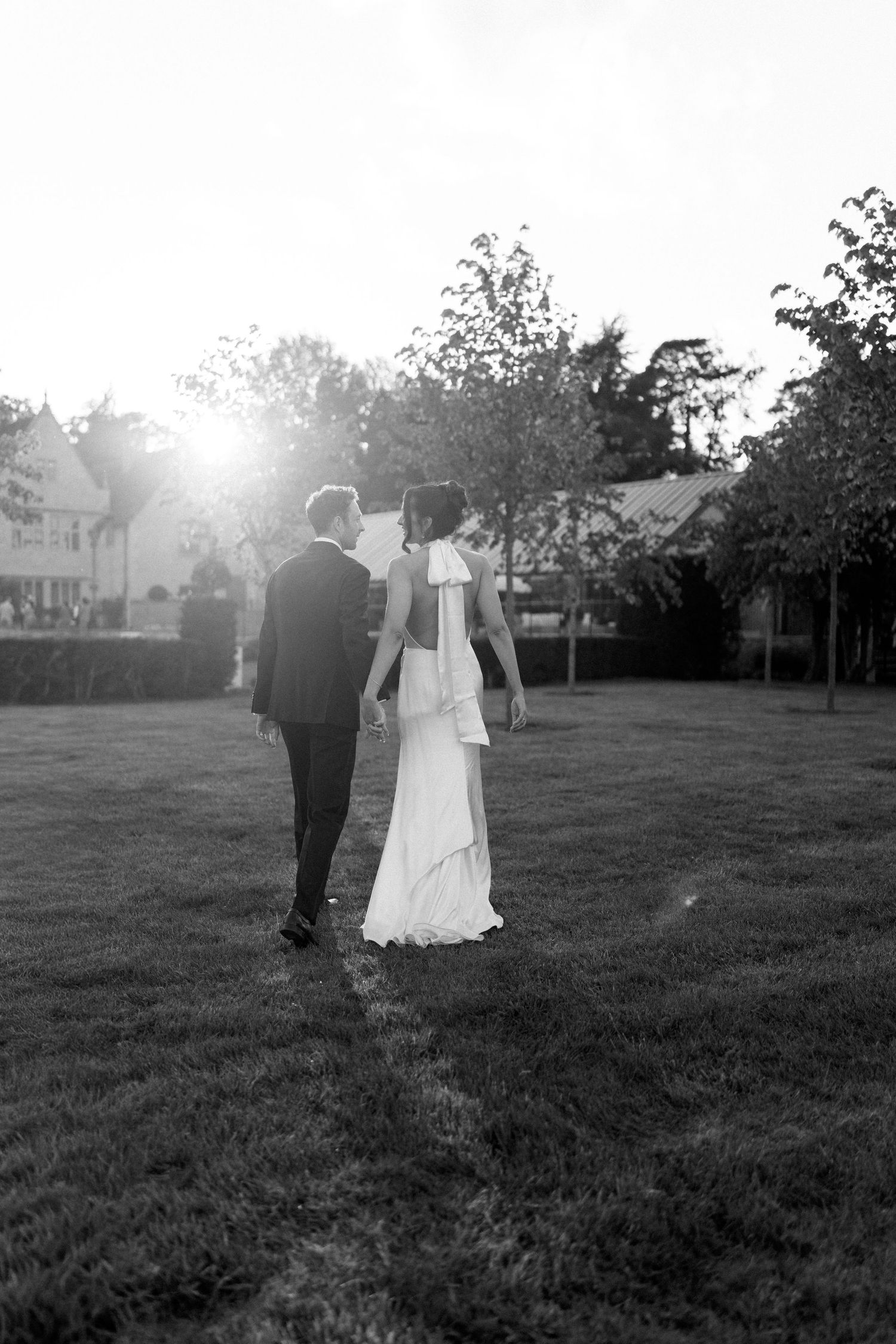 Bride and groom walk hand in hand across lawn in stunning backlit black and white sunset portrait.