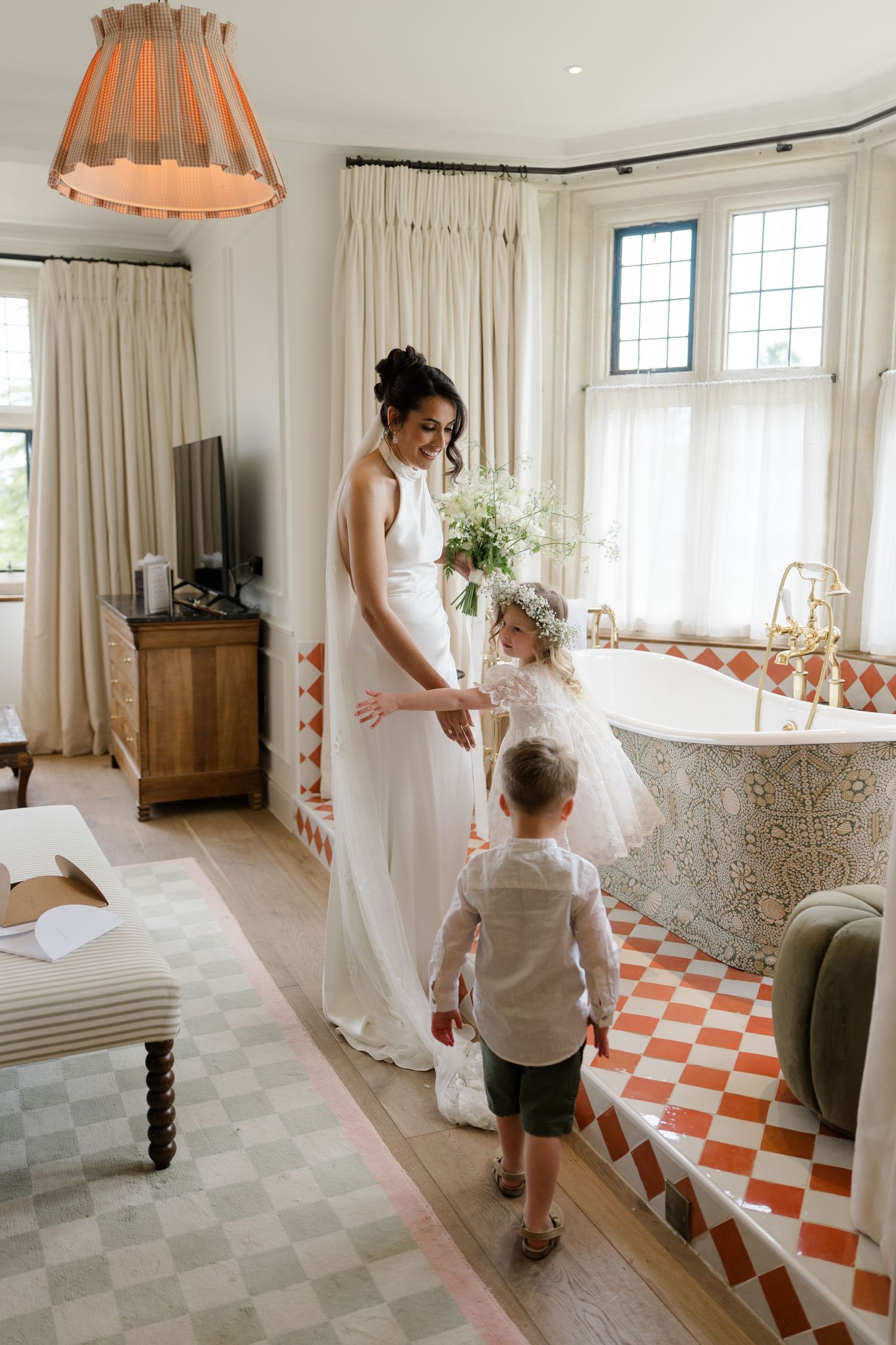 Bride in white halter gown with flower girl and page boy in ornate tiled bathroom.