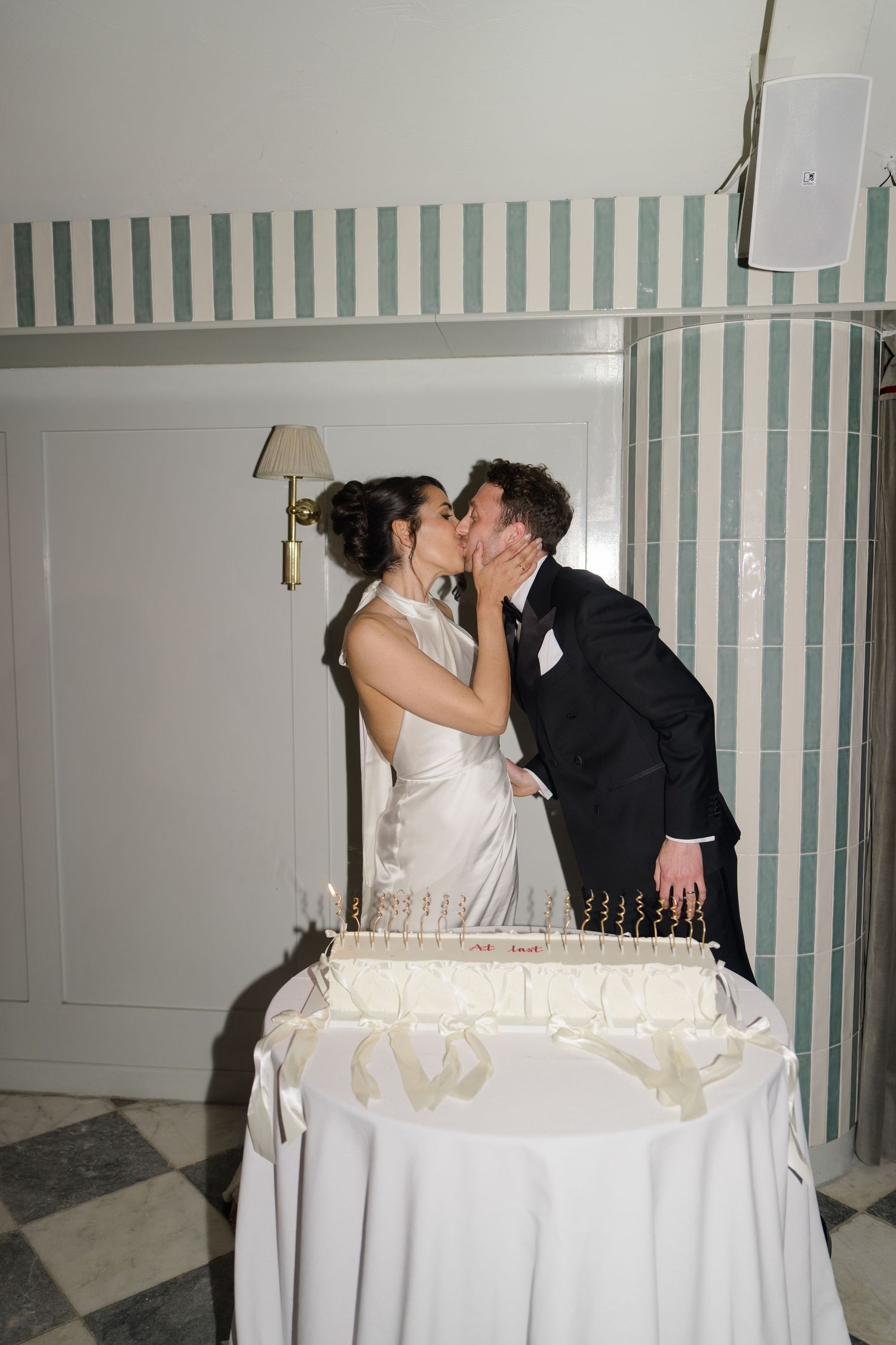 Bride and groom share romantic kiss over wedding cake at striped wall reception venue.