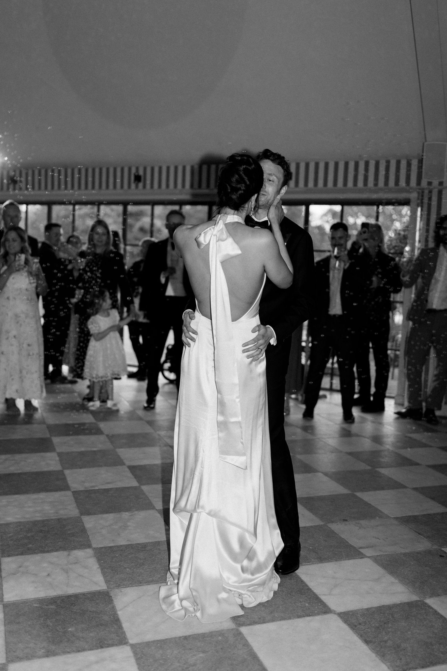 Bride and groom share first dance on checkered floor surrounded by guests.