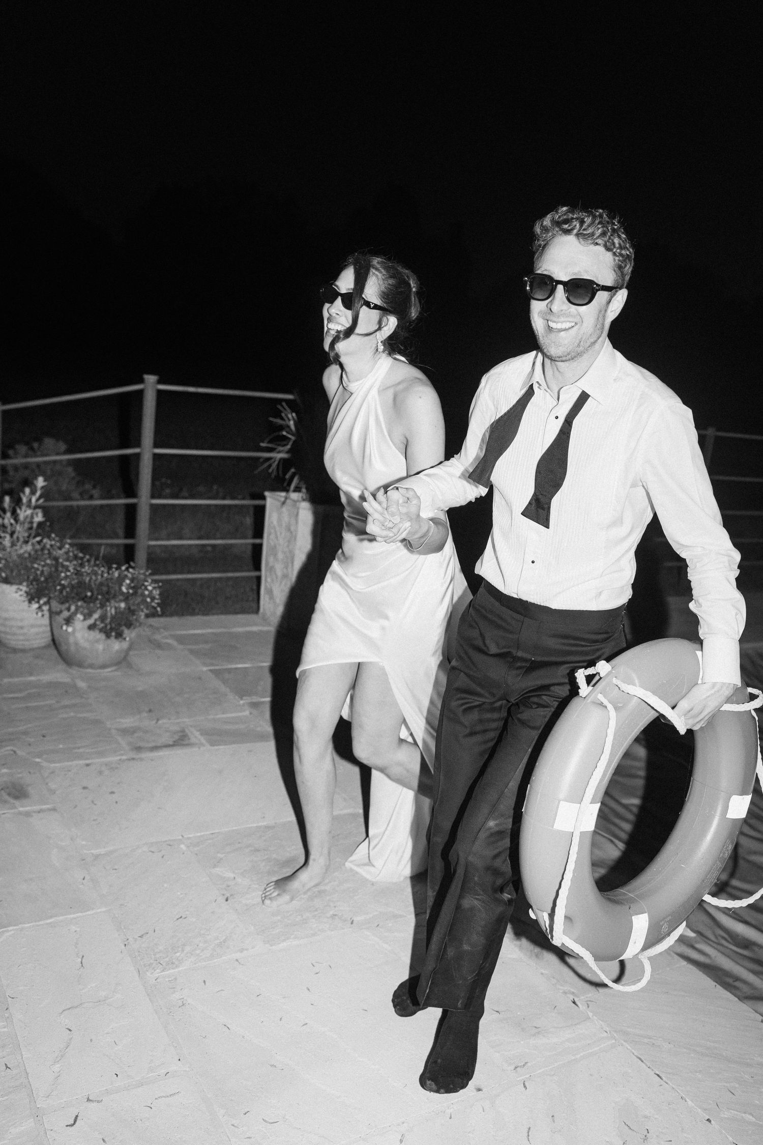 Black and white photo of smiling newlyweds in sunglasses, groom holding life ring at night.
