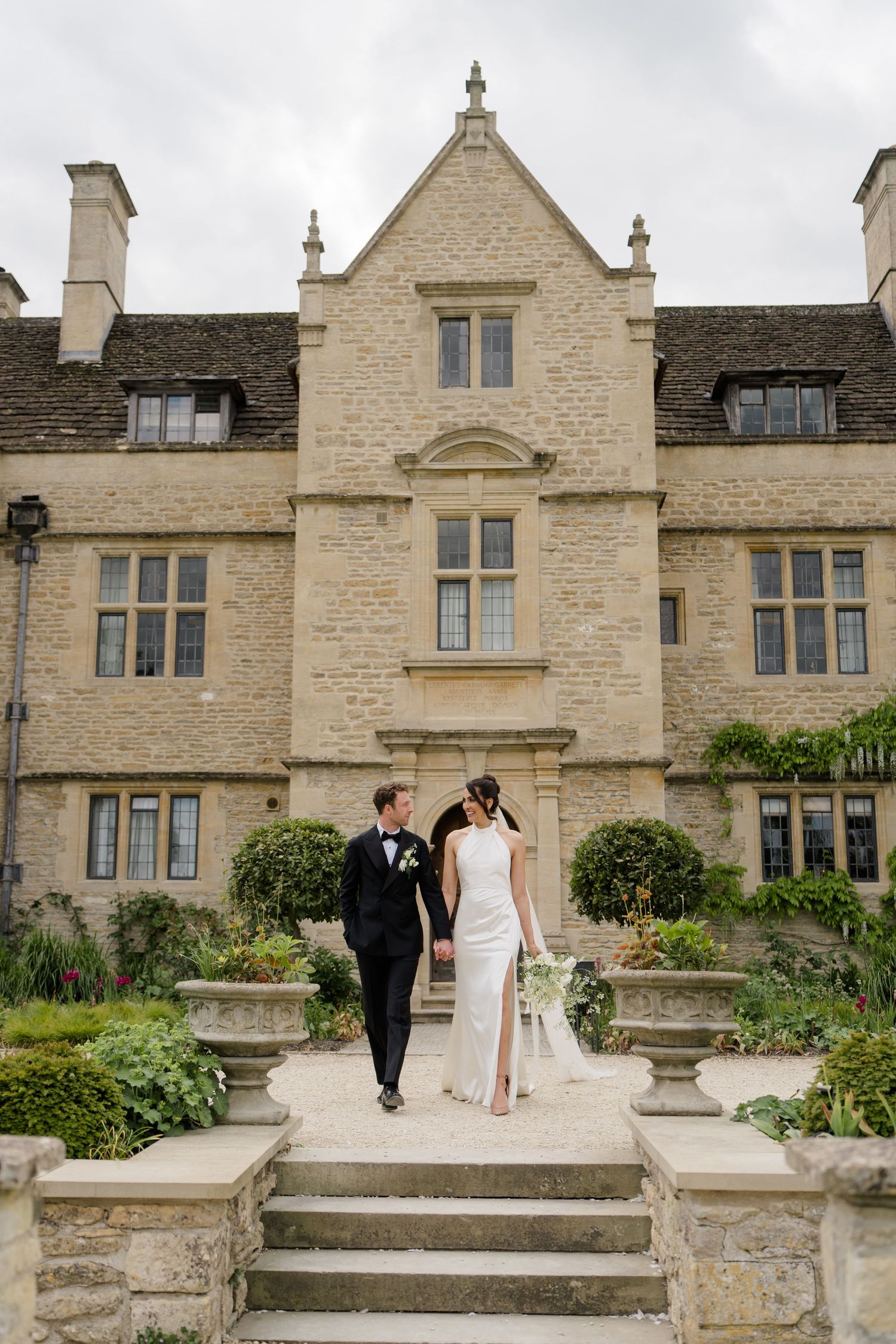 Newlyweds descend stone steps hand in hand outside a stunning Cotswolds manor house wedding venue.