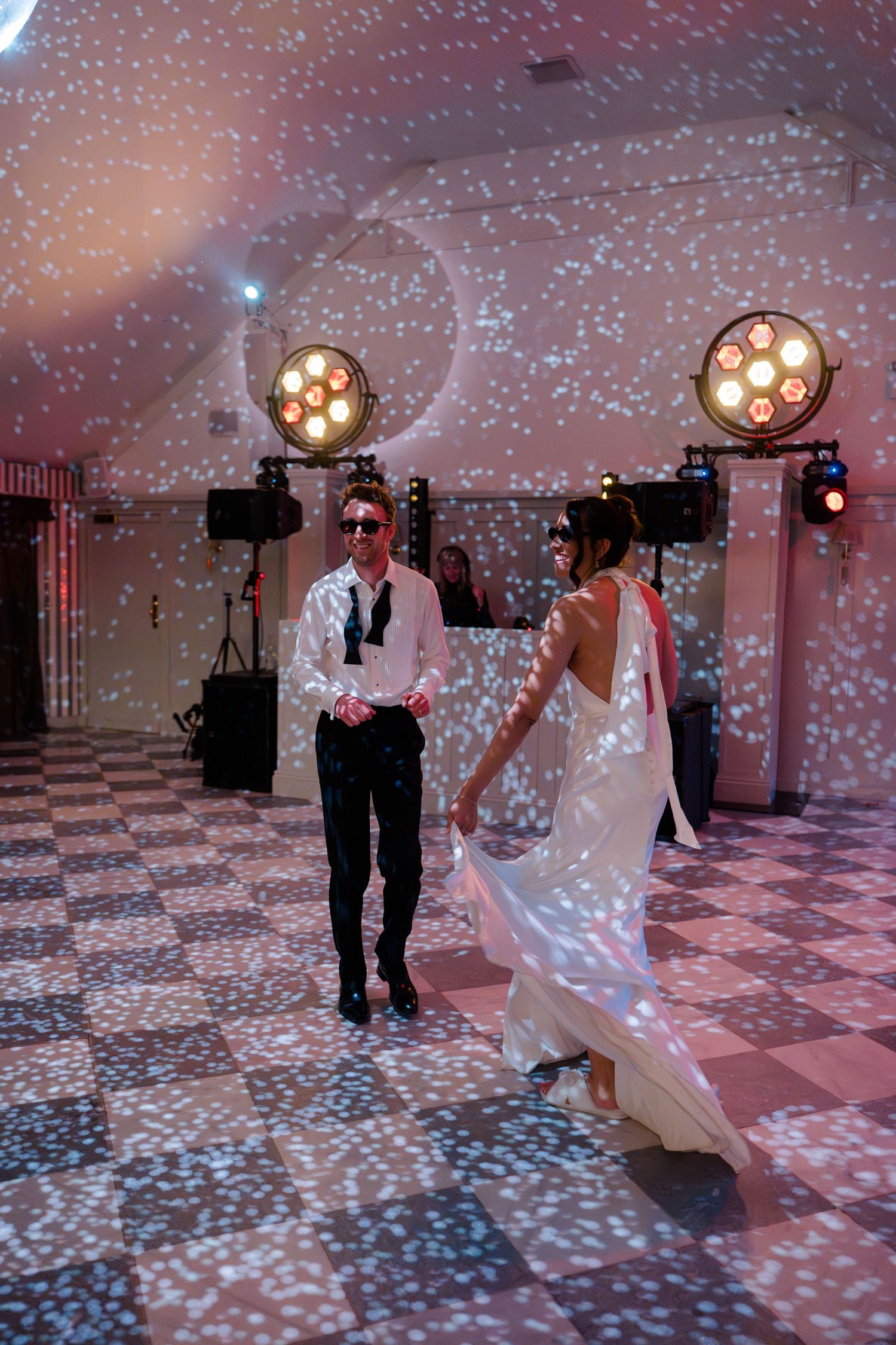 Bride in flowing white gown and groom dancing under sparkling lights on wedding dance floor.