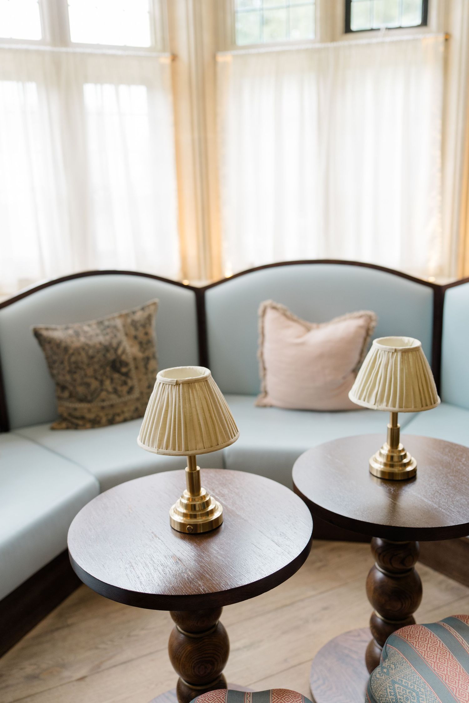 Elegant bay window seating area with light blue curved sofa, decorative pillows, and two brass table lamps on dark wood side tables.