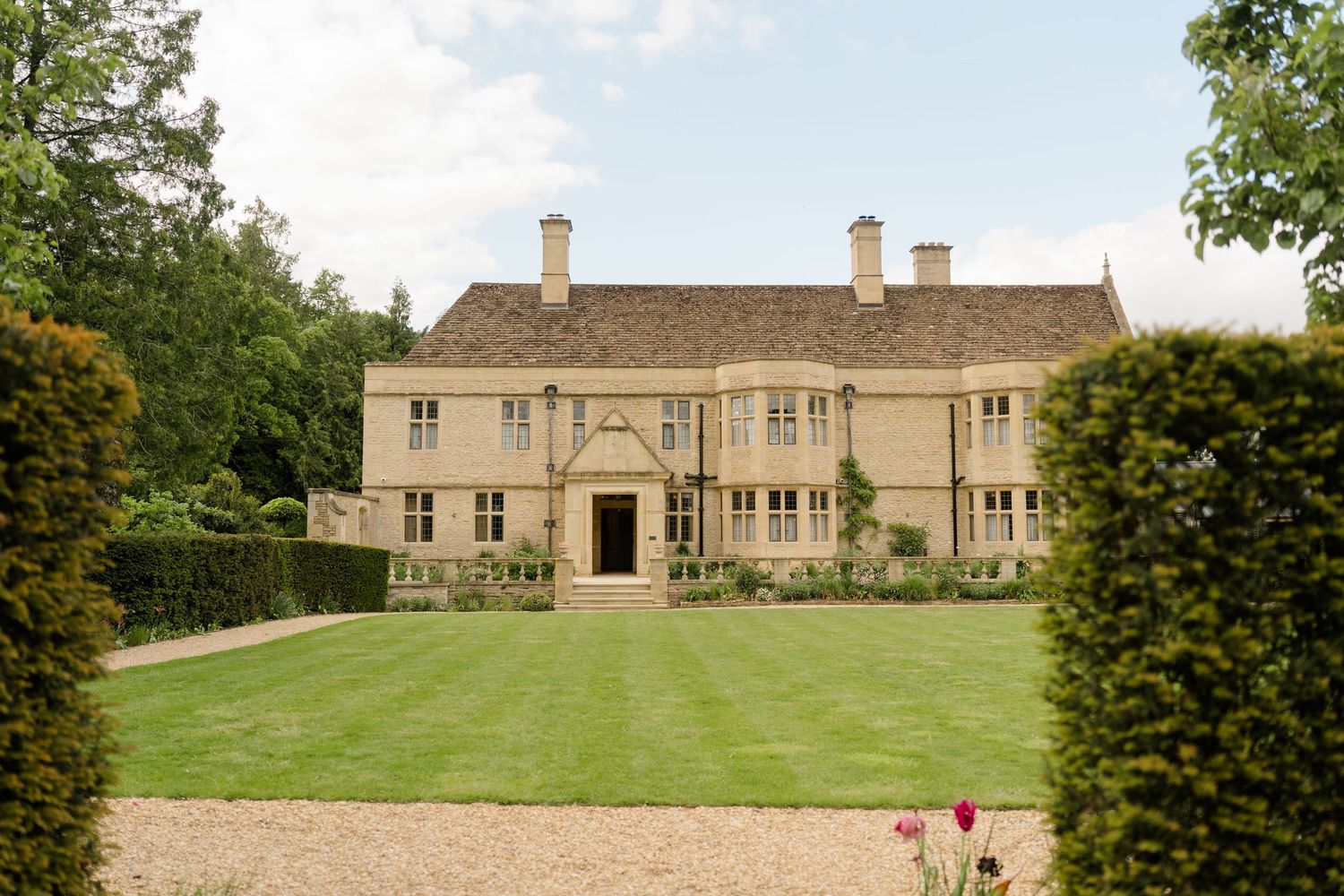 A grand Cotswold stone manor house with manicured lawns, topiary hedges, and climbing roses on a sunny day.
