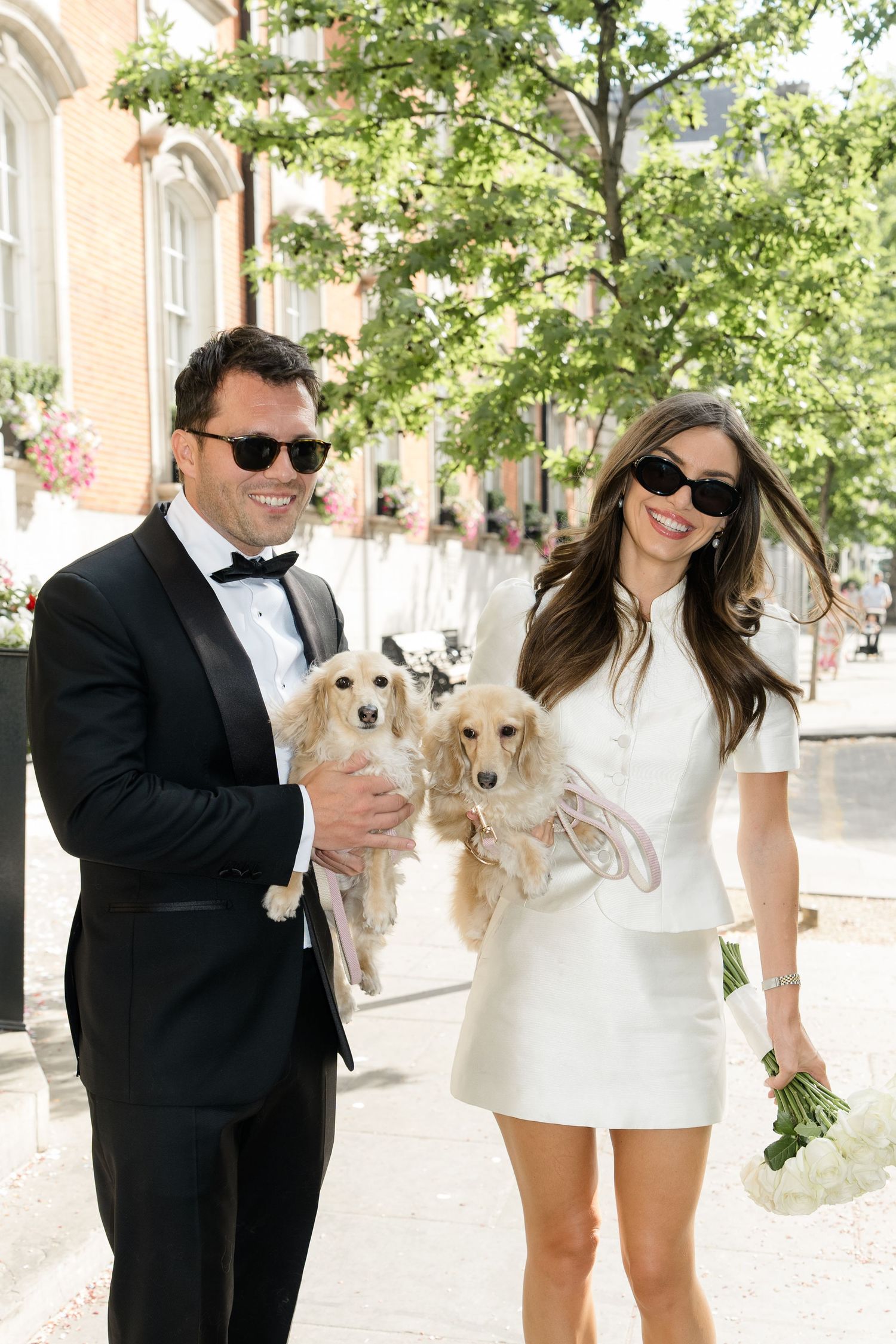 Smiling couple in wedding attire holding two cocker spaniels outdoors.
