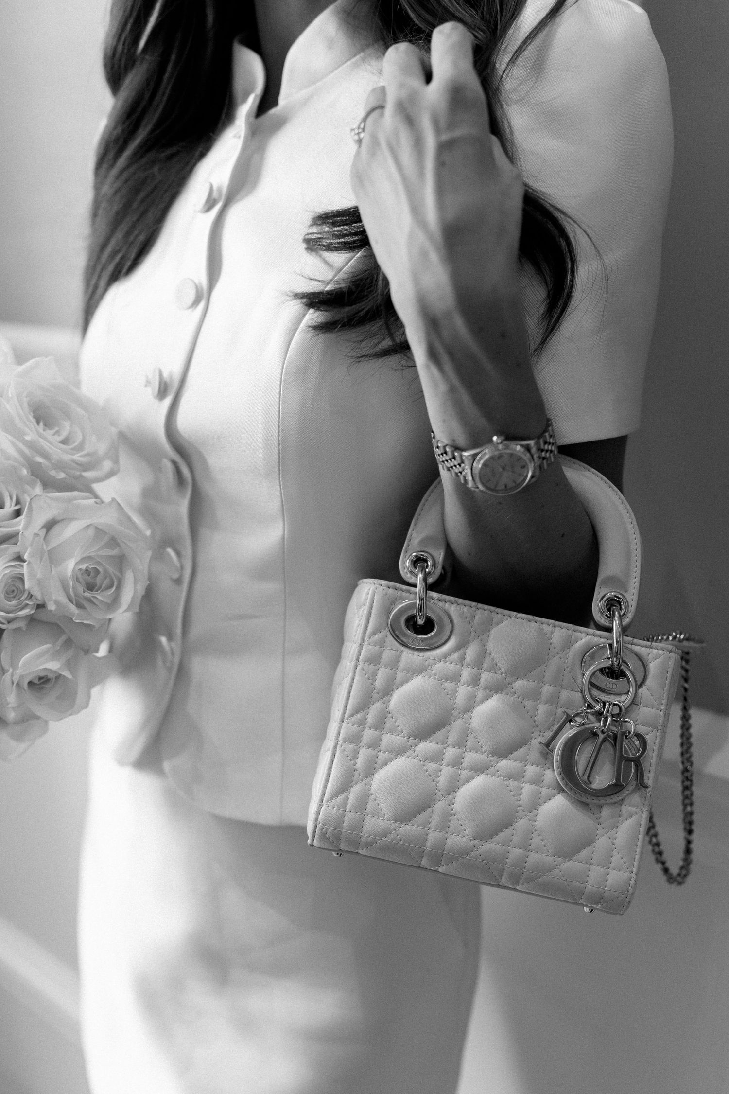 Black and white close-up of bride in white outfit holding Lady Dior handbag and white rose bouquet.
